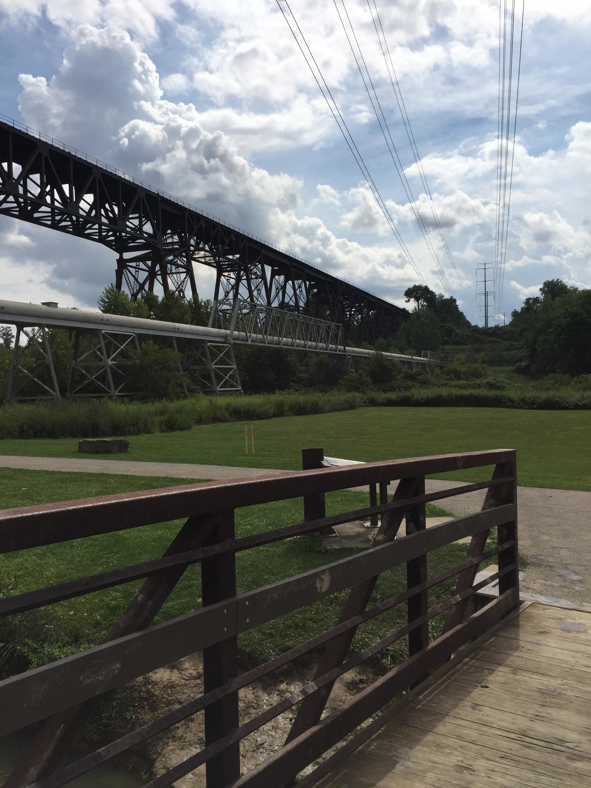 A view of a large metal railway bridge from below, with a pipe running parallel beneath it. The scene includes a grassy area and a pathway in the foreground, framed by a wooden railing. The sky is partly cloudy, with a mix of blue and white clouds. Electric power lines run above the bridge. Ohio And Erie Canal mountain bike trail.