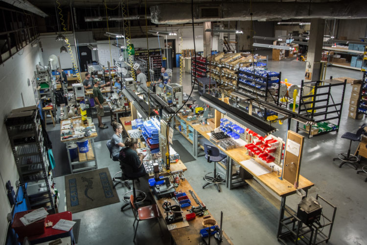 A high-angle view of a busy manufacturing workspace, featuring several workers engaged in various tasks. The area is filled with workbenches, tools, and storage bins organized in vibrant colors. Industrial lighting hangs overhead, illuminating the space, which showcases a blend of machinery and manual labor in a well-structured environment.
