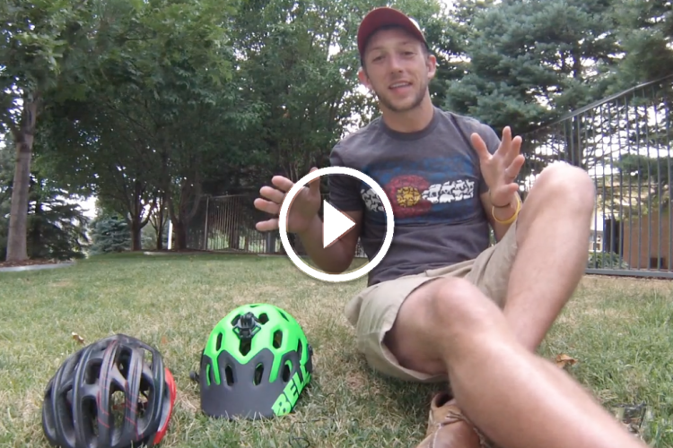 A person is sitting on the grass with two bicycle helmets in front of them—a black one and a bright green one. They are wearing a casual shirt and shorts, and appear to be explaining something while gesturing with their hands. Trees and a fence are visible in the background. A play button overlay indicates that this is a video.