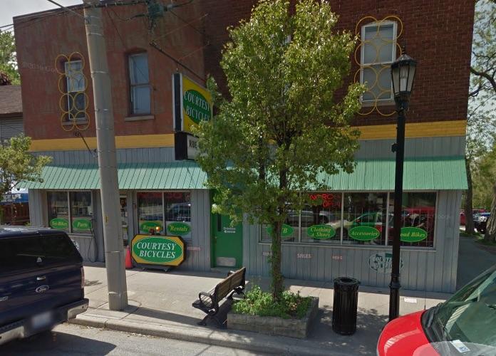 Front view of Courtesy Bicycles shop, featuring a green awning and large windows displaying various bicycle-related services. A small tree is in front, with a bench nearby, and a street lamp illuminates the area. The building has a brick upper section with a yellow trim.