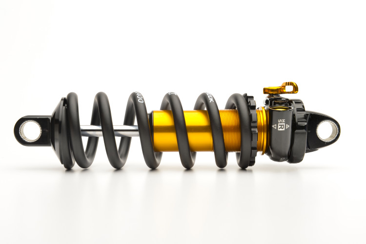 A high-quality image of a shock absorber, featuring a prominent black coil spring surrounding a vibrant orange shock body. The top of the shock shows an adjustable mechanism in black with a small golden toggle, and the ends have eyelets for installation. The background is plain white, emphasizing the design and details of the shock absorber.