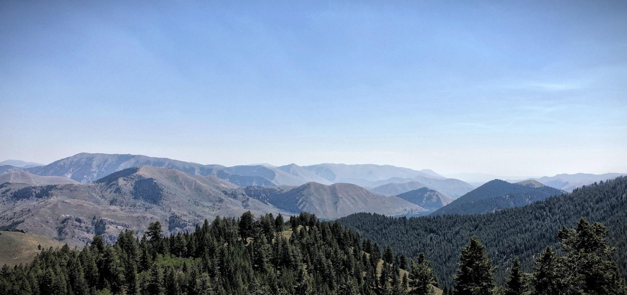 A panoramic view of a mountainous landscape featuring rolling hills covered in patches of evergreen trees under a clear blue sky. The image captures the layers of mountains fading into the distance, with varying shades of green and brown across the terrain. Bald Mountain Bike Park mountain bike trail.