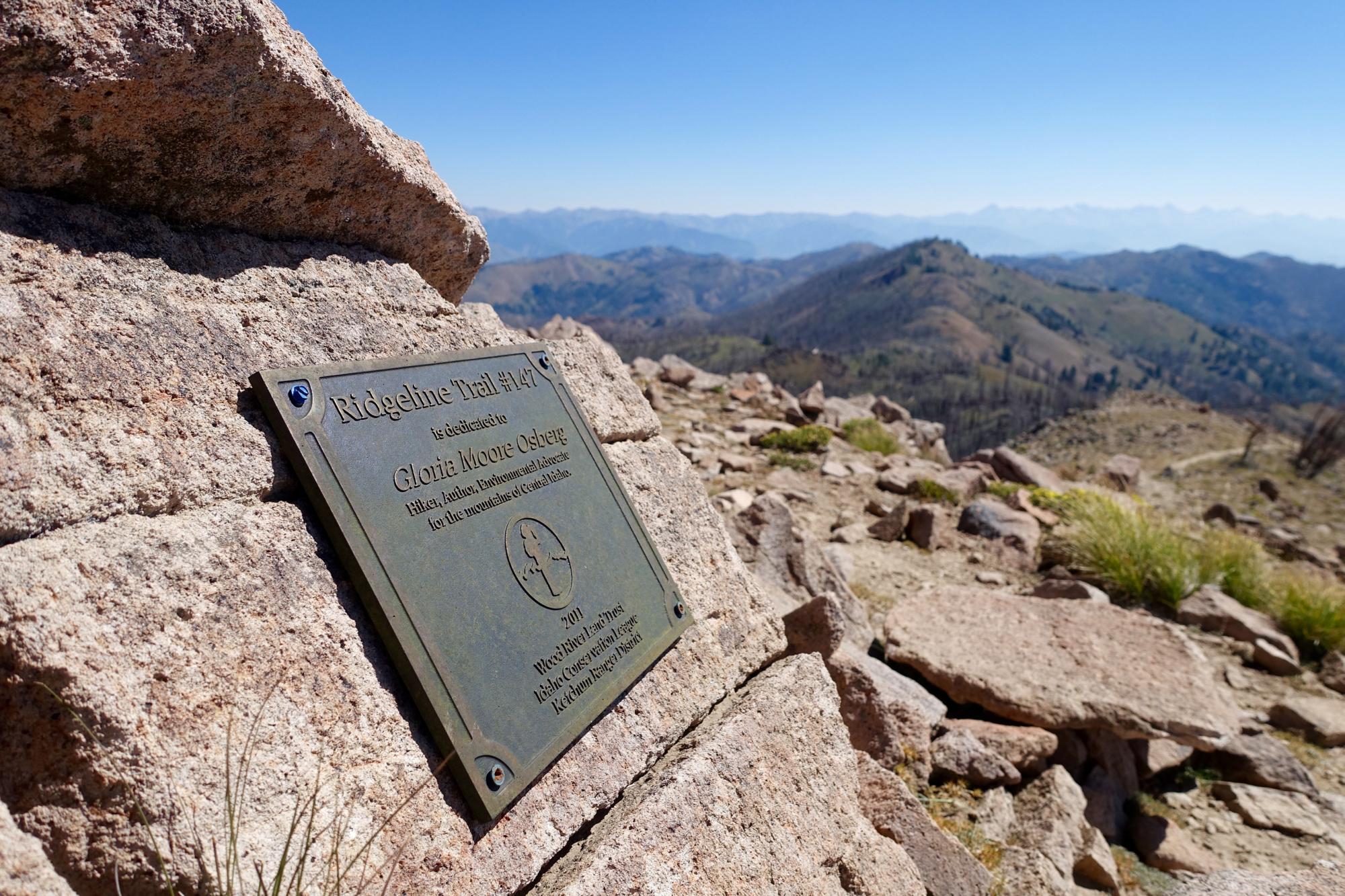 A bronze plaque mounted on a rocky surface, dedicated to Gloria Moore Osberg, marking Ridgeline Trail #147. The background features rolling mountains under a clear blue sky. Osberg's Ridgeline Trail mountain bike trail.