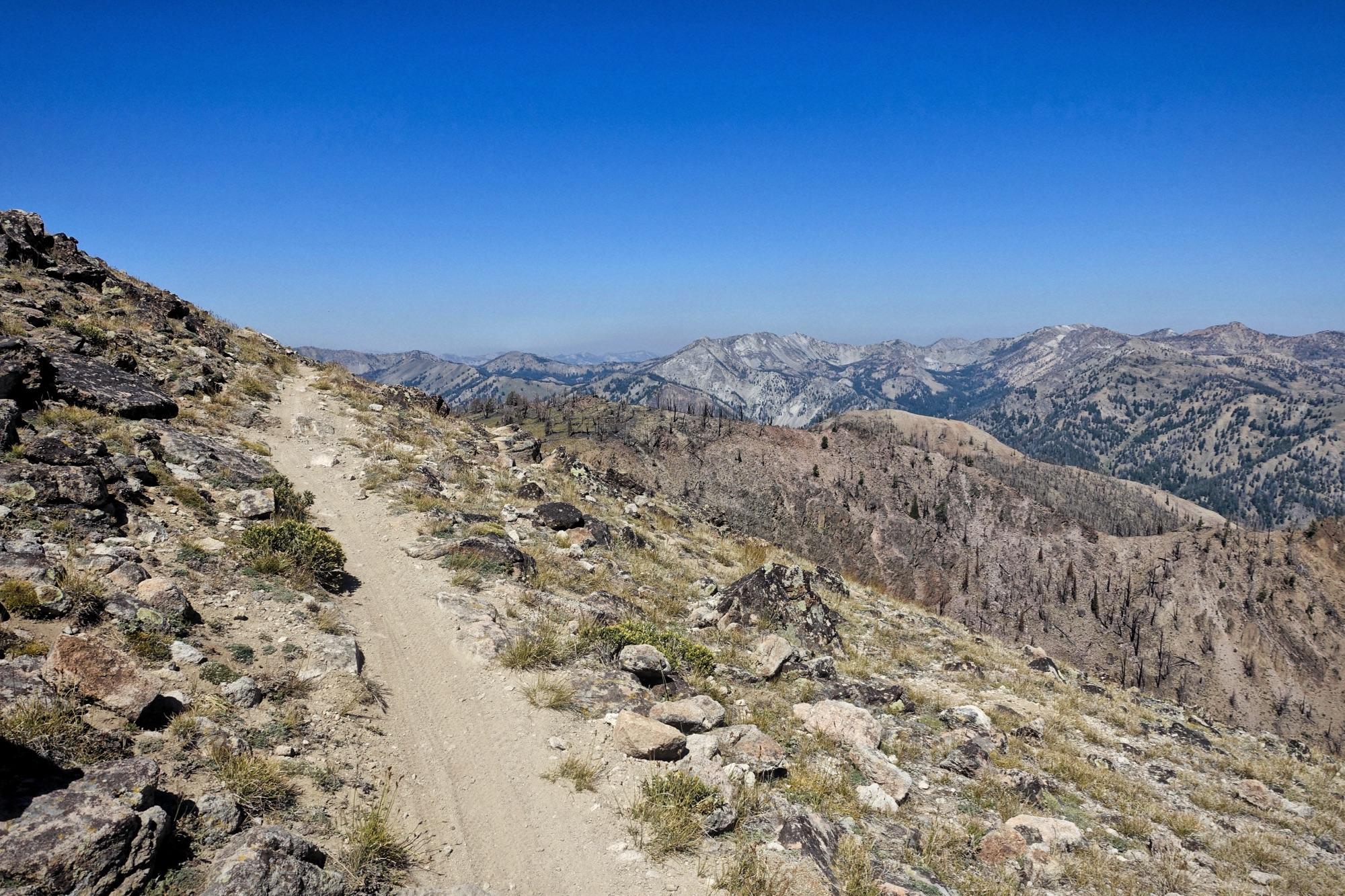A narrow dirt trail winds along a rocky mountain ridge, surrounded by sparse vegetation and scattered stones. In the background, a range of mountains stretches across the horizon under a clear blue sky. The scene conveys a sense of remote wilderness and natural beauty. Osberg's Ridgeline Trail mountain bike trail.