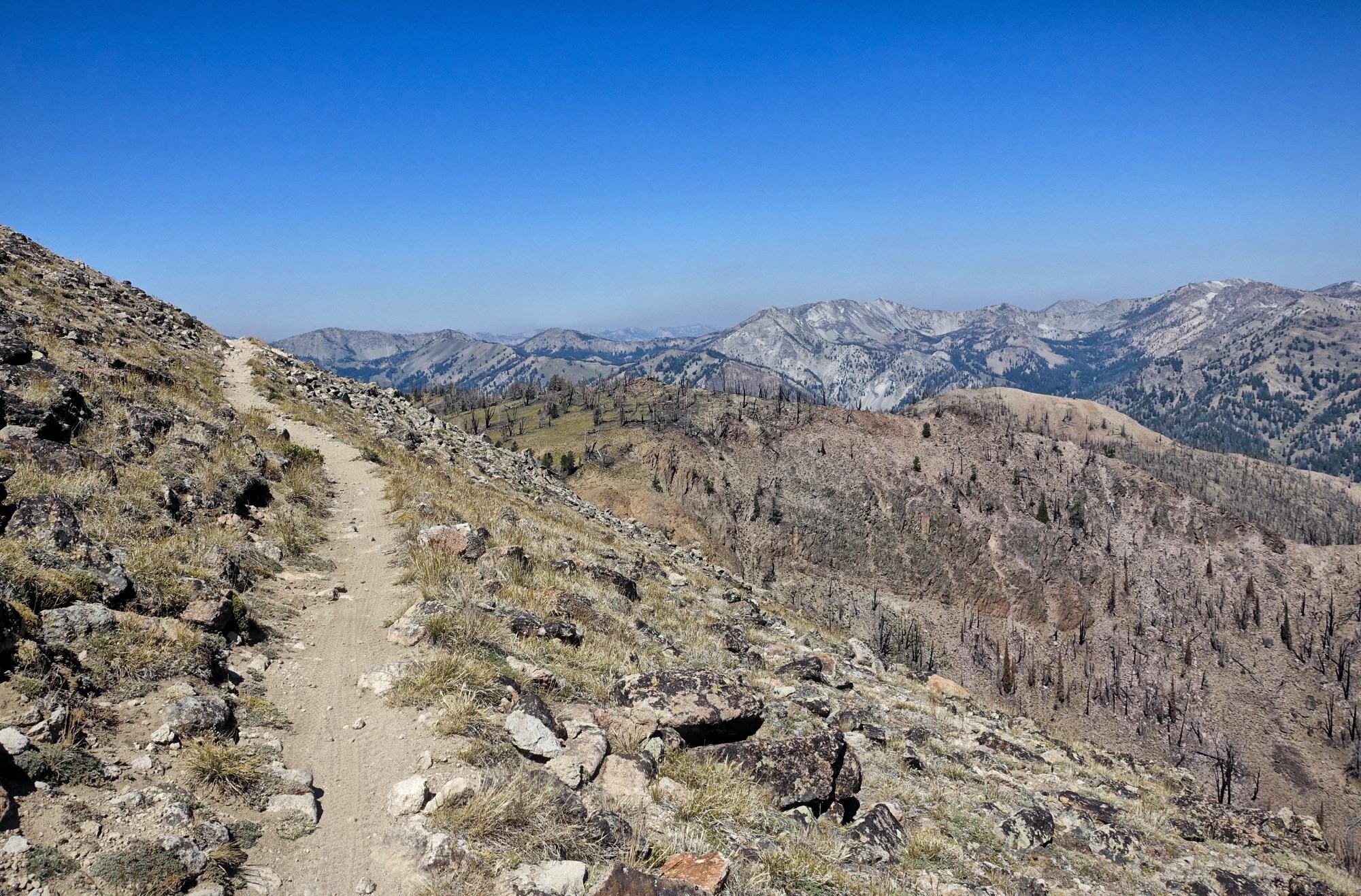 A narrow dirt hiking trail winds along a rocky hillside, offering expansive views of distant mountains under a clear blue sky. The landscape features patches of grass and scattered rocks, with signs of past tree growth amid the rugged terrain. Osberg's Ridgeline Trail mountain bike trail.