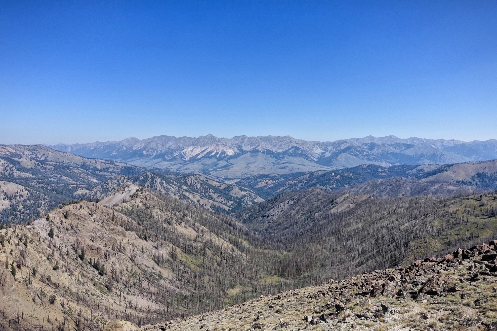 A panoramic view of a mountainous landscape under a clear blue sky. The scene includes rolling hills, ridges, and distant peaks, some with rocky formations and patches of greenery, interspersed with areas of scorched trees, indicating past forest fires. Osberg's Ridgeline Trail mountain bike trail.