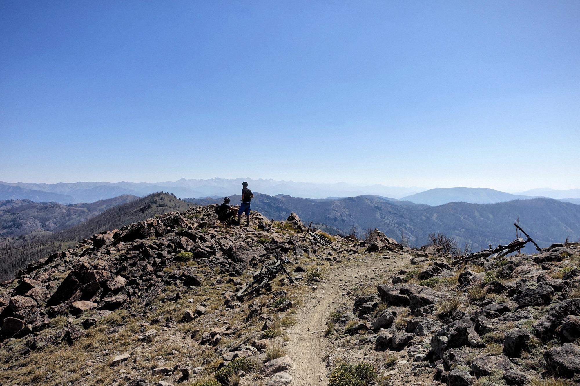 A hiker and a dog stand on a rocky mountain peak, overlooking a vast landscape of rolling hills and distant mountains under a clear blue sky. A dirt trail winds through the rocky terrain, while a bike leans against the rocks nearby. Osberg's Ridgeline Trail mountain bike trail.