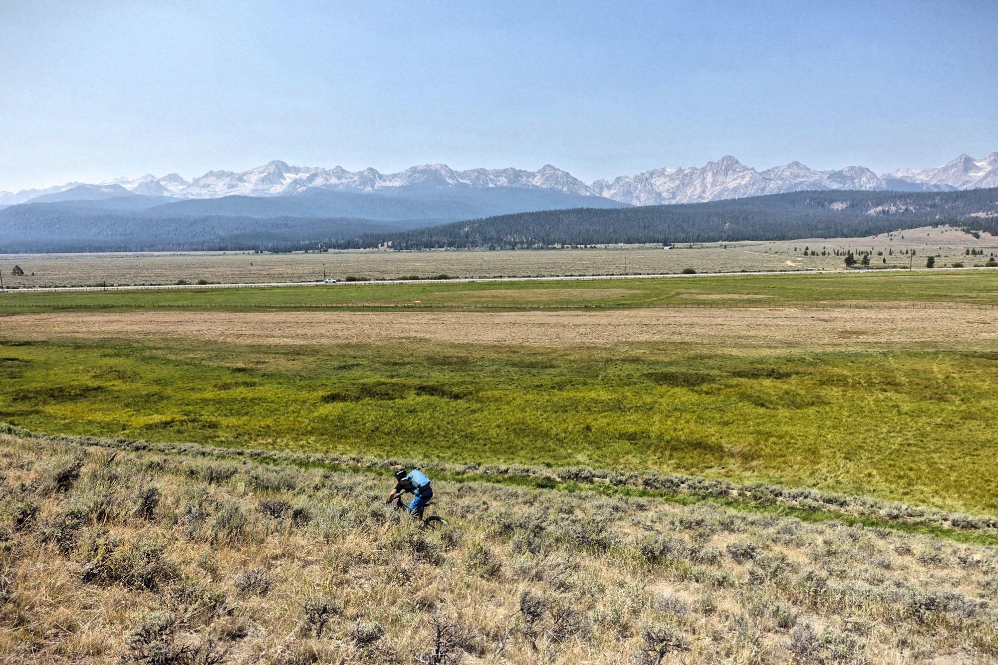 A cyclist navigating a grassy hillside with a panoramic view of distant mountains under a clear sky. The landscape features open fields with varying shades of green and brown, stretching towards the horizon. Williams Creek mountain bike trail.