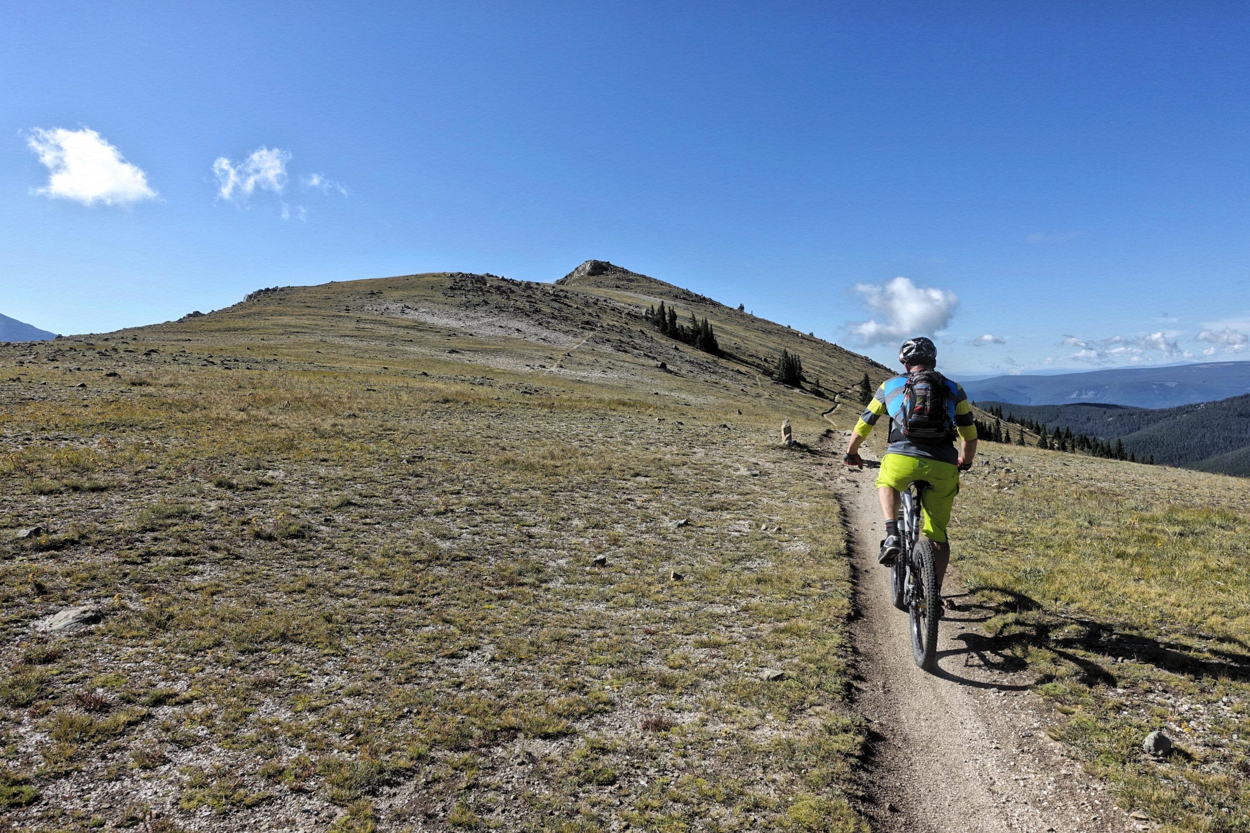 A mountain biker riding along a dirt trail on a sunny day, surrounded by grassy hills and distant mountains under a blue sky with a few clouds. The biker is wearing bright yellow shorts and a striped shirt, with a backpack and helmet. Monarch Crest Trail mountain bike trail.