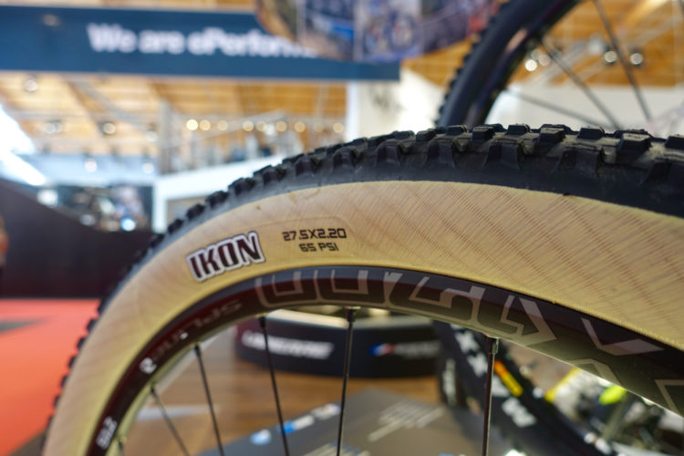 Close-up of a mountain bike tire showing the tread pattern. The tire is labeled "IKON" with specifications indicating a size of 27.5 x 2.20 and a maximum pressure of 65 PSI. The background features a blurred exhibition space with a sign that reads "We are ePerformance."