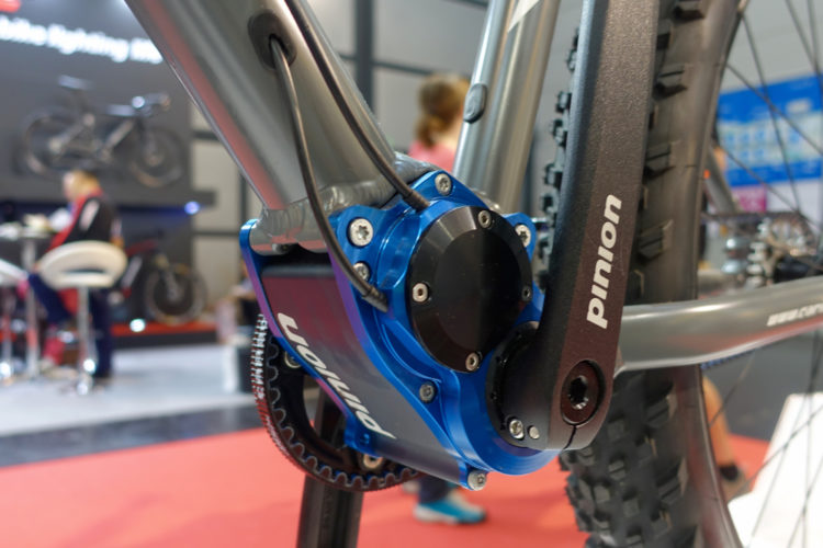Close-up view of a bicycle's mid-section, showcasing the blue gear system and crank arm labeled "Pinion." The background features a blurred image of people at a bike exhibit, with a red carpet floor.