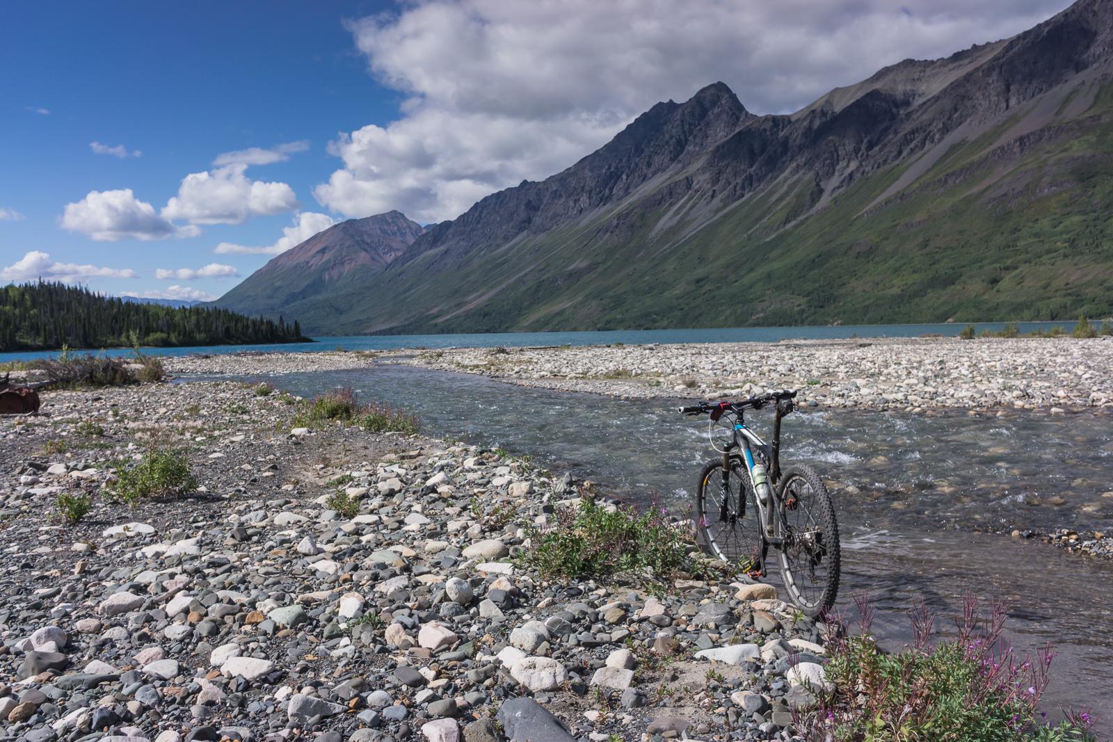 A mountain bike parked on a rocky riverbank, surrounded by pebbles and wildflowers, with a backdrop of steep mountains and a clear blue sky dotted with clouds. The river flows gently beside the bike, while a lush green forest can be seen on the opposite bank. Cottonwood Trail mountain bike trail.