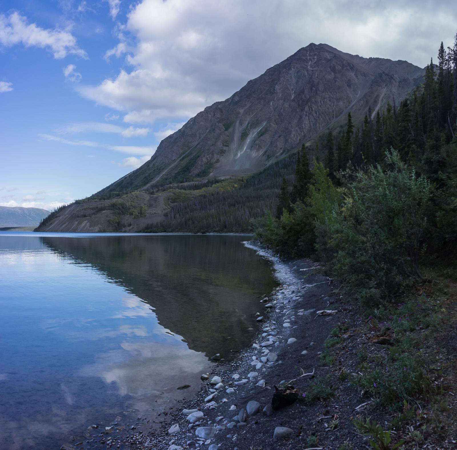 A serene lake reflecting a mountainous landscape under a partly cloudy sky. The foreground features a rocky shore lined with smooth stones and lush greenery, while the backdrop showcases a towering mountain covered with trees and rocky slopes. Cottonwood Trail mountain bike trail.