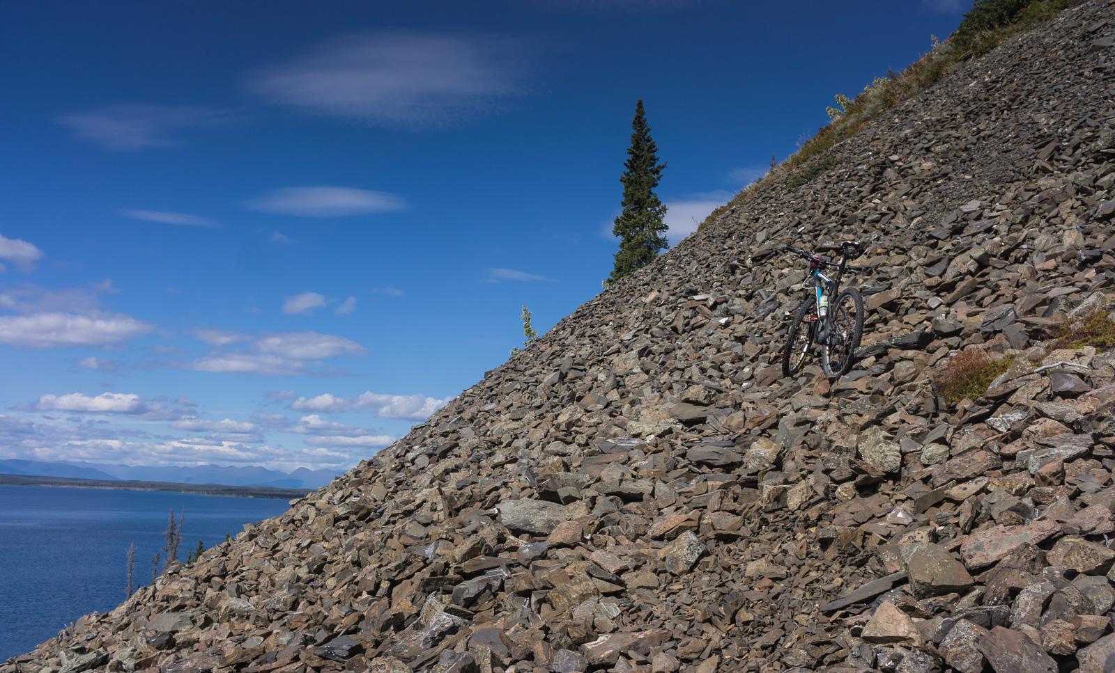 A mountain bike resting on a rocky slope overlooking a serene lake, with a clear blue sky and scattered clouds in the background. Cottonwood Trail mountain bike trail.