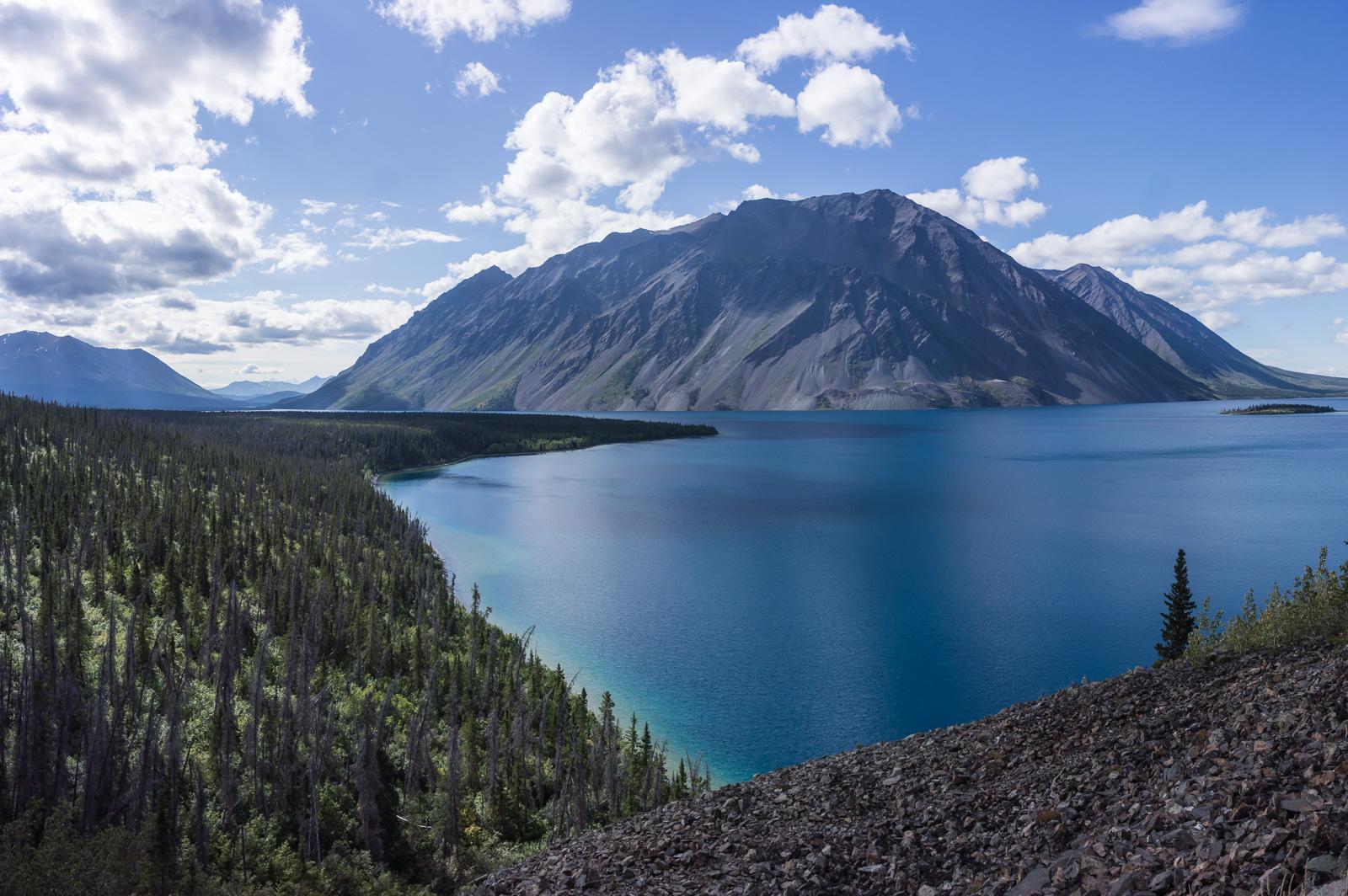 A panoramic view of a serene lake surrounded by lush green forests and towering mountains under a partly cloudy sky. The lake