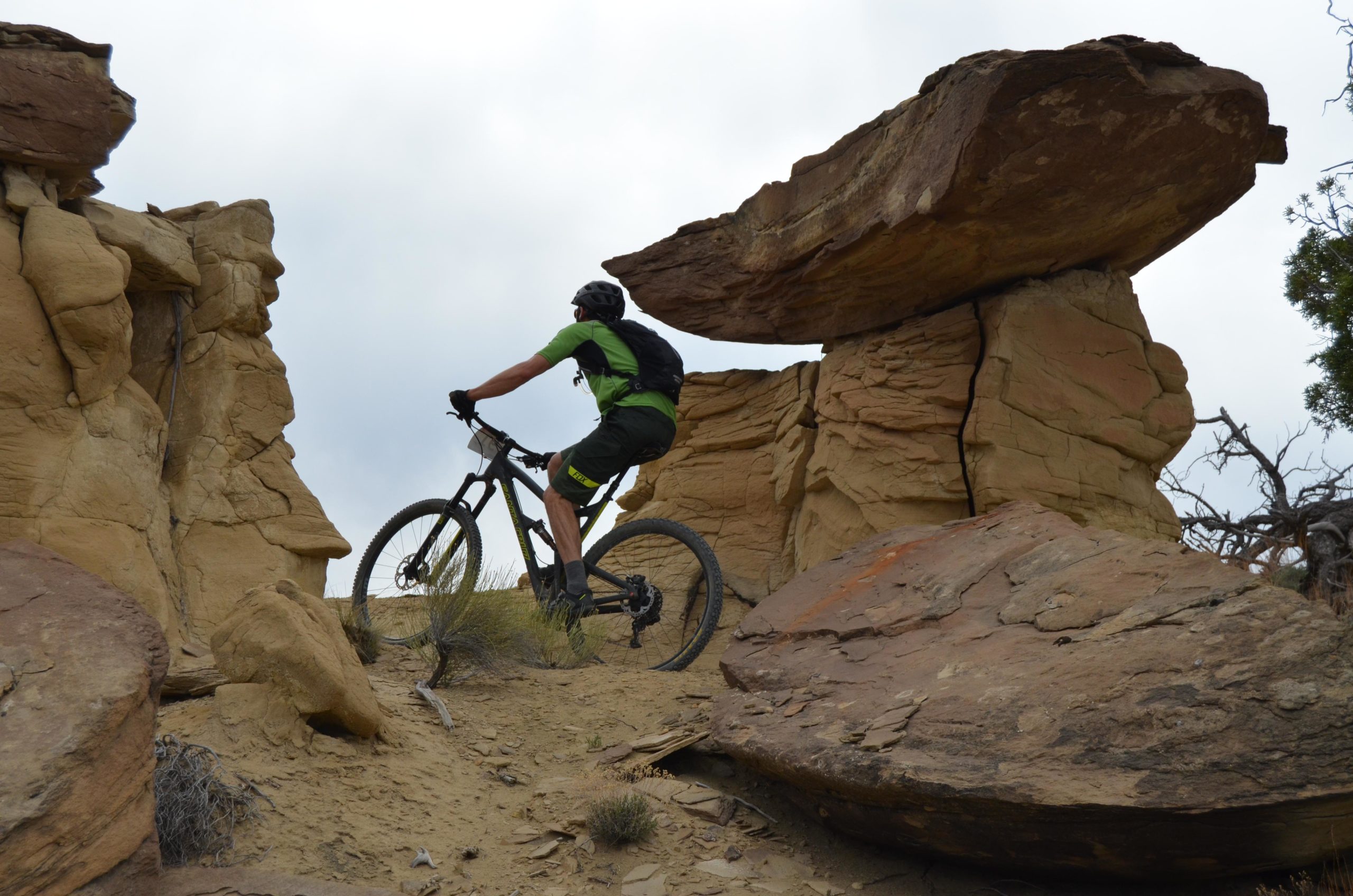 A mountain biker navigating a rocky terrain with large boulders, surrounded by sandy soil and sparse vegetation under a cloudy sky. High Desert Trail System mountain bike trail.