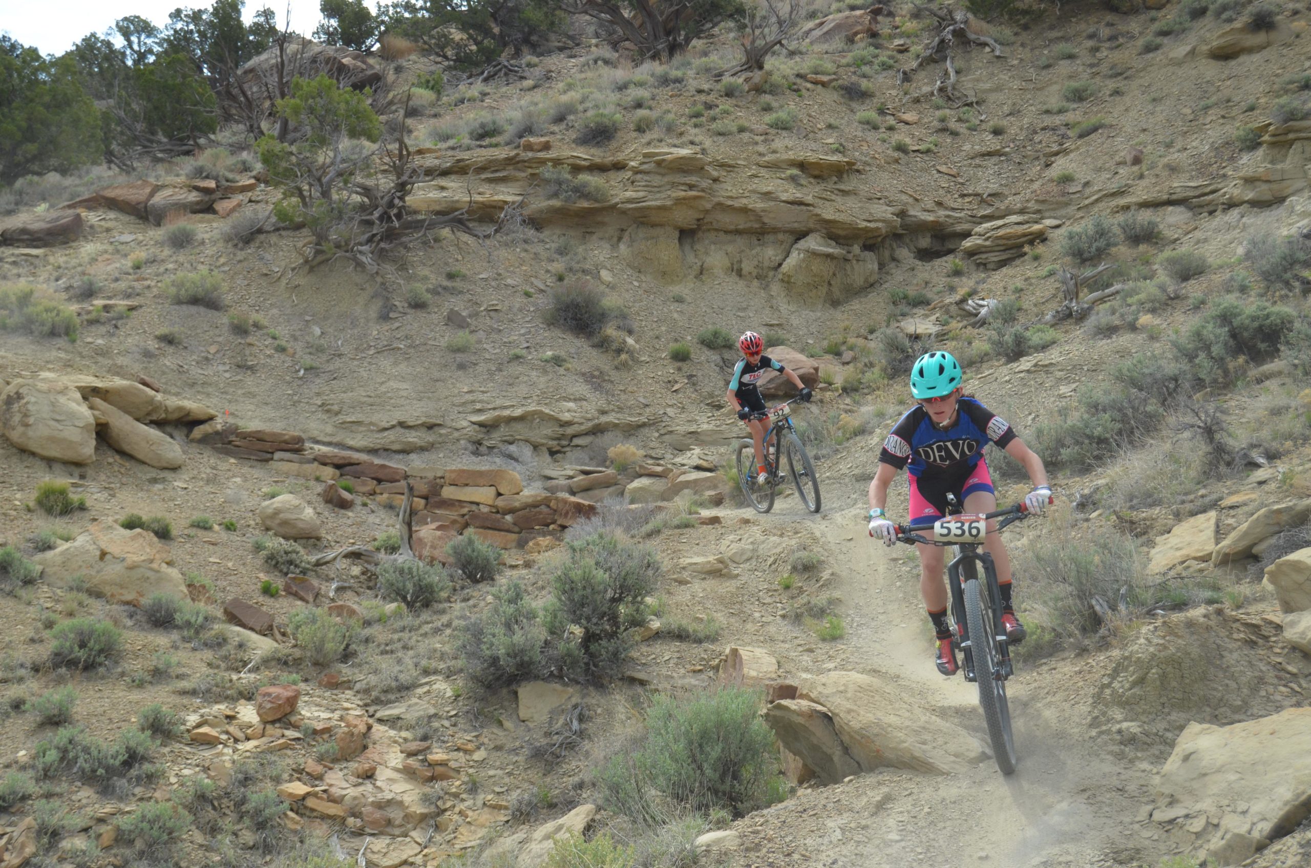Two mountain bikers navigate a dirt trail in a rocky, arid landscape. One rider is wearing a teal helmet and black jersey with colorful shorts, focused on maneuvering downhill, while the other rider, in a black jersey and red helmet, is climbing the trail behind. Surrounding them are shrubs and rock formations typical of a dry outdoor setting. High Desert Trail System mountain bike trail.