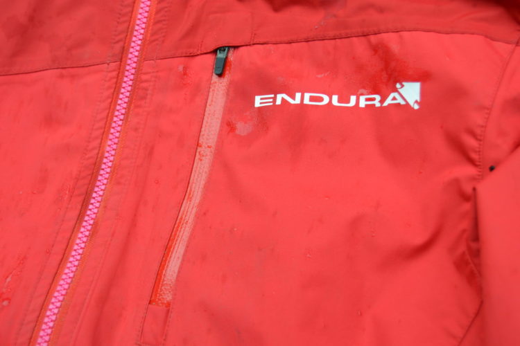 Close-up of a bright red waterproof jacket featuring a pink zipper and the logo "ENDURA" on the chest area. The fabric shows signs of moisture with droplets visible on the surface.