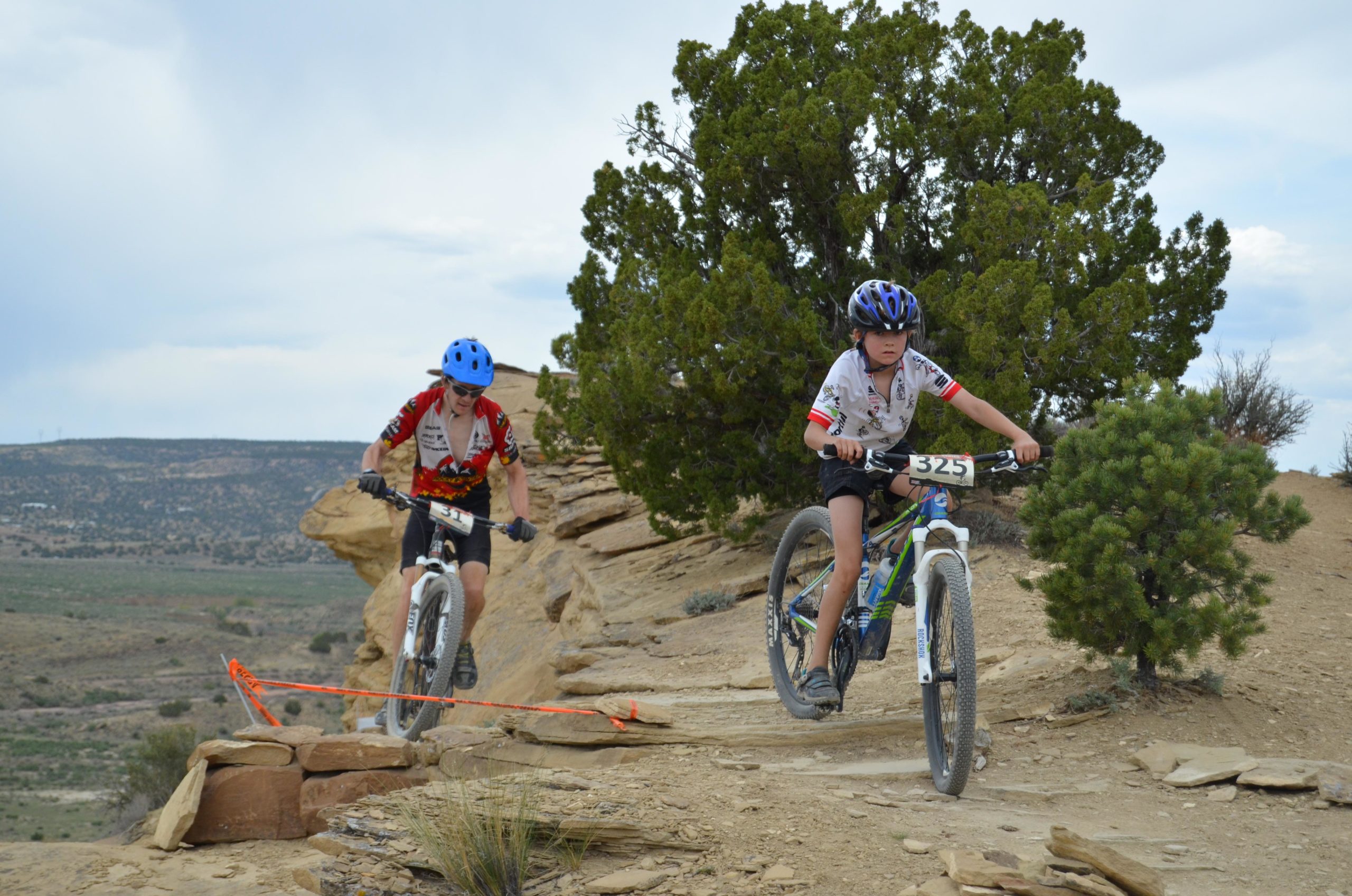 Two young cyclists compete in a mountain bike race on a rocky trail surrounded by sparse vegetation. One boy, wearing a blue helmet and a red and black jersey, navigates a challenging section of the path, while the other, with a black helmet and a white jersey, rides with focus. The landscape features rolling hills and a cloudy sky in the background. High Desert Trail System mountain bike trail.