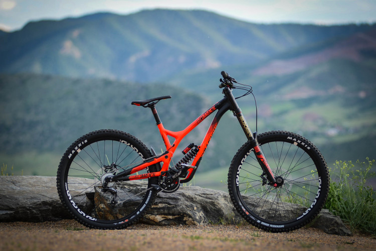 A vibrant orange and black mountain bike is positioned on a rocky surface, with a backdrop of lush green mountains under a partly cloudy sky. The bike features wide tires suitable for rugged terrain and a sturdy frame designed for off-road riding.