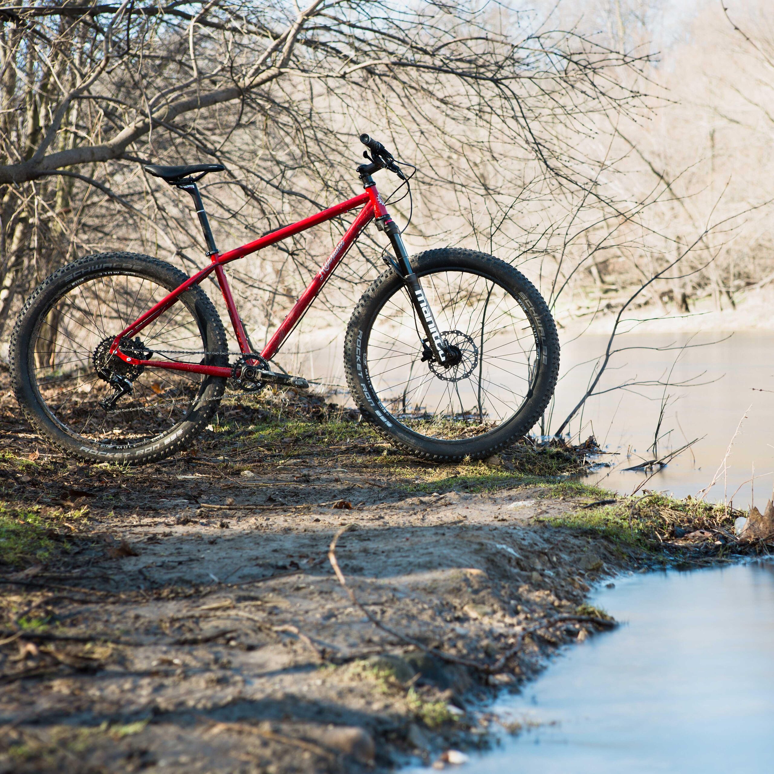 A red mountain bike leaning on a gravel and dirt trail by a calm river, surrounded by bare trees in a natural setting.