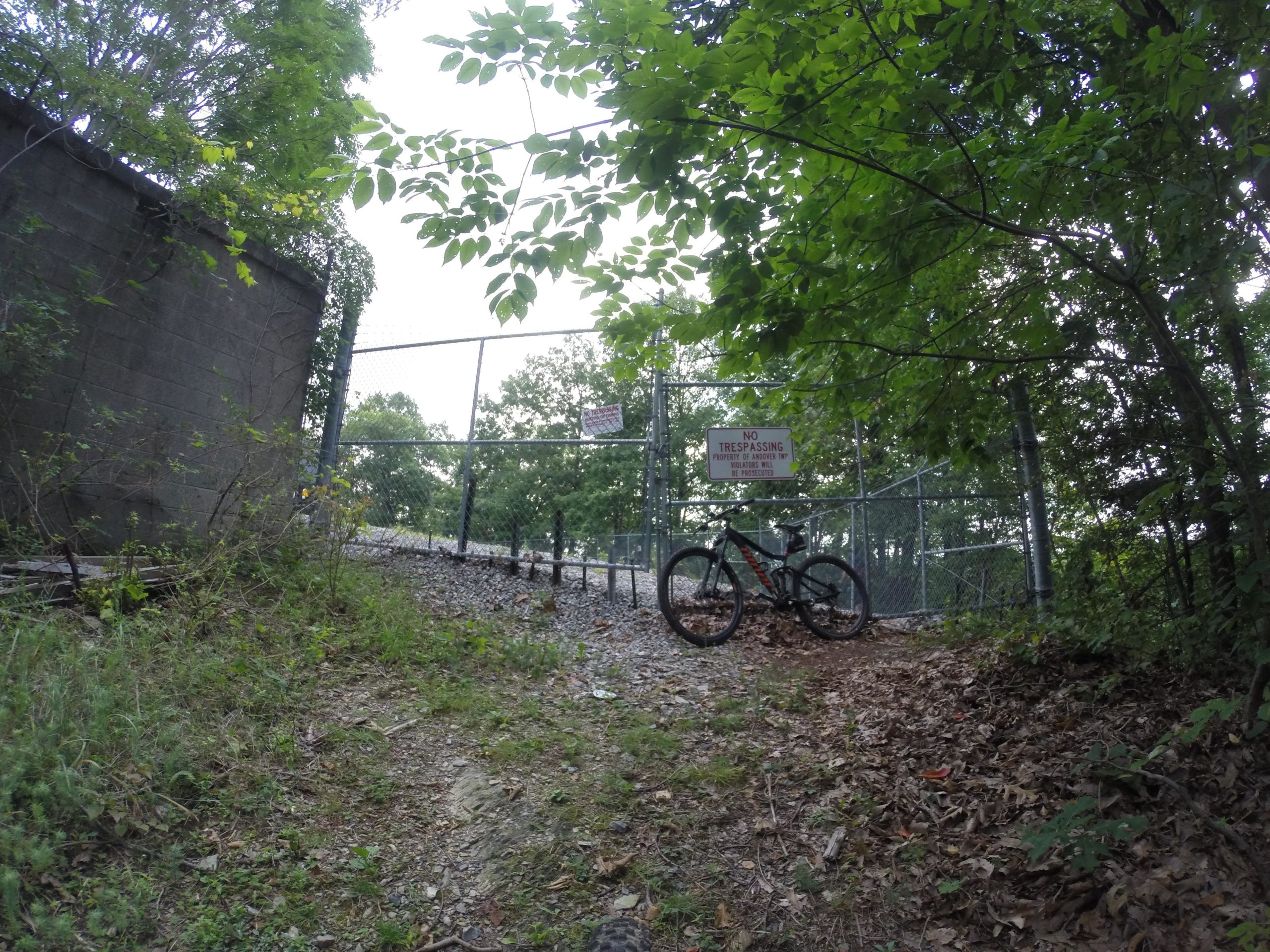 A mountain bike is resting on a gravel path surrounded by dense foliage, near a chain-link fence with a "No Trespassing" sign posted. The view captures a secluded area that appears to lead into the woods. Kittatiny Valley State Park mountain bike trail.