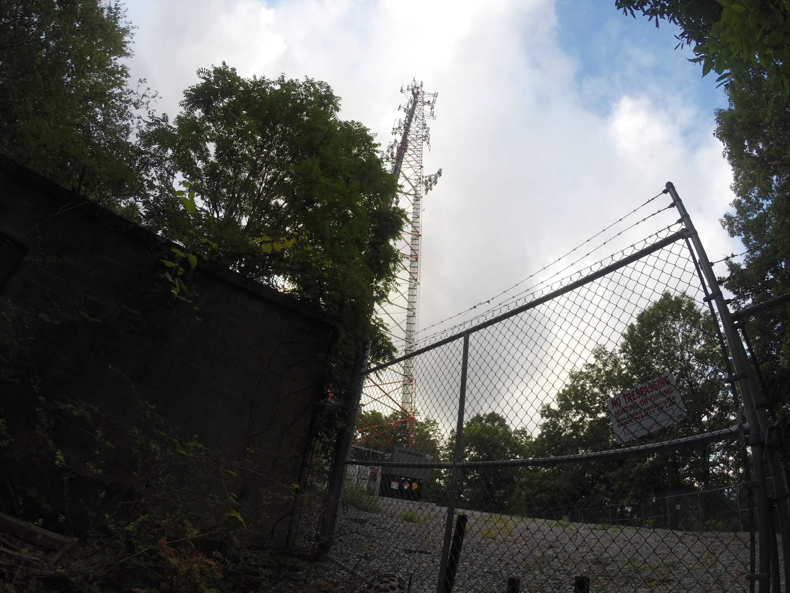 A view through a chain-link fence of a tall communications tower surrounded by trees, with a cloudy sky in the background. A "No Trespassing" sign is visible on the fence. Kittatiny Valley State Park mountain bike trail.