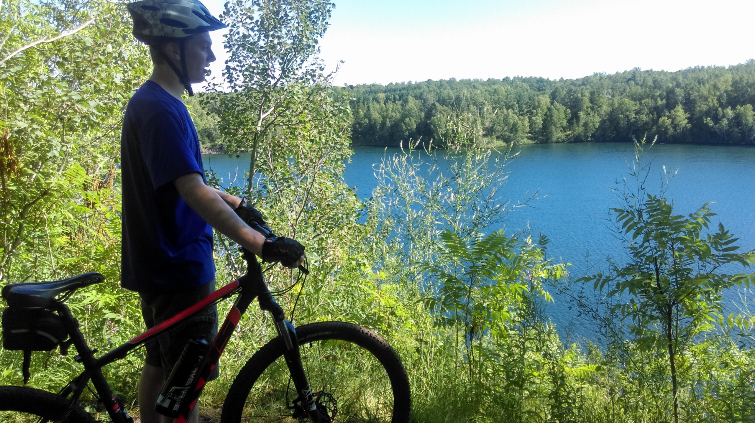 A person in a blue shirt and helmet stands next to a mountain bike, gazing at a calm, blue lake surrounded by lush greenery and trees on a sunny day. Cuyuna Lakes mountain bike trail.
