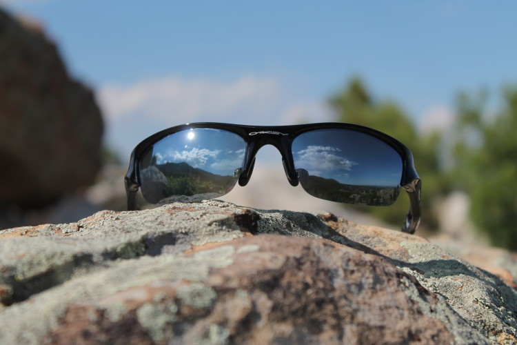 A pair of black Oakley sunglasses resting on a textured rock, reflecting a blue sky with fluffy clouds and distant mountains in the lenses.