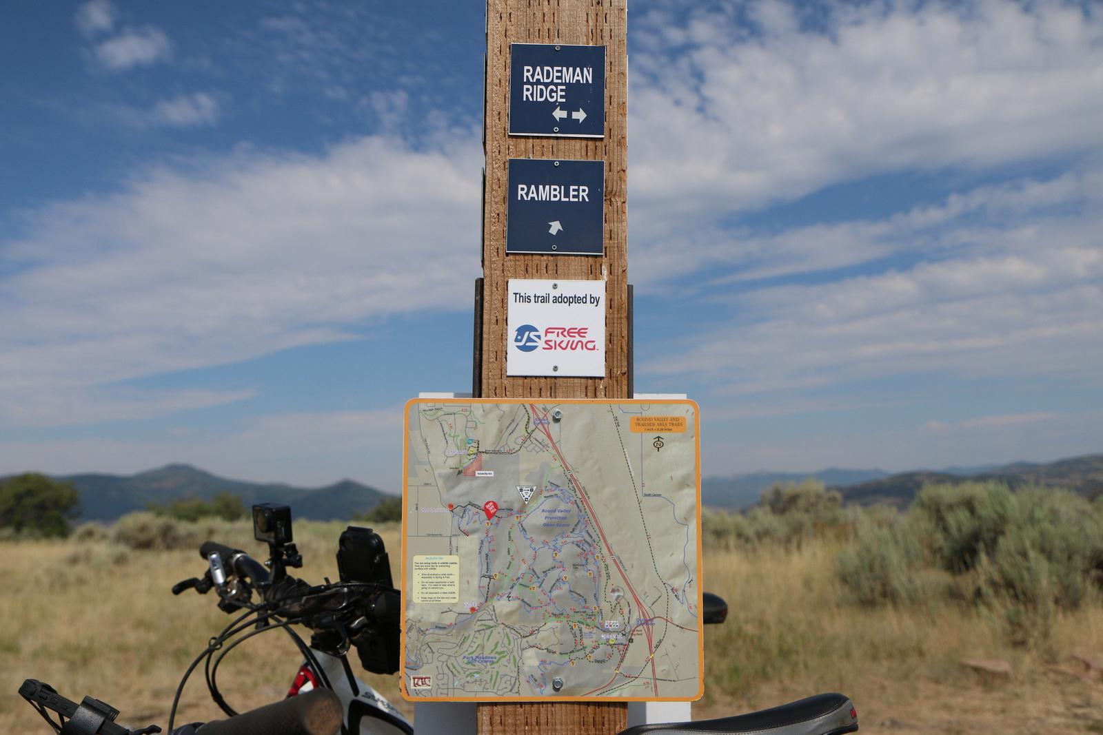 A wooden trail sign displaying directions for "Rademan Ridge" and "Rambler" trails, along with a trail map. The sign indicates that the trail is adopted by "Free Skiing." In the foreground, part of a bike is visible, and the background features a landscape of hills and clear skies. Round Valley mountain bike trail.