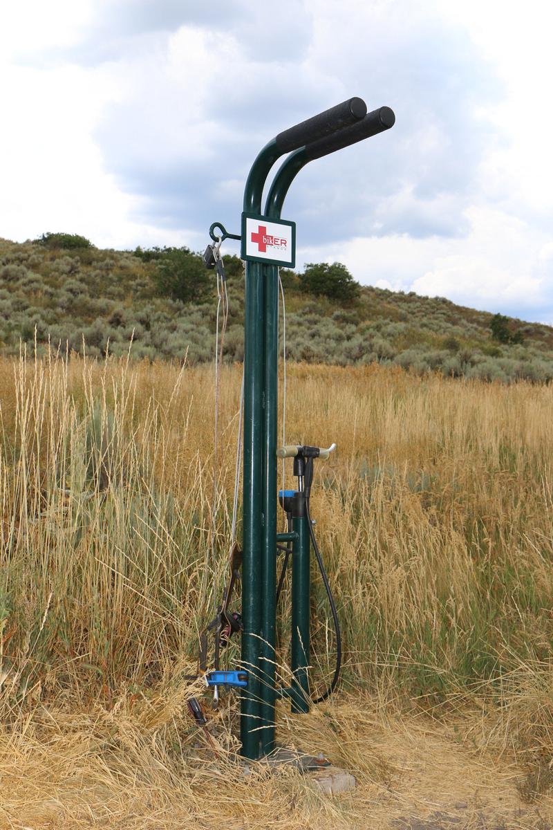 A bike repair station in a grassy area, featuring a green metal frame with two handlebars and various tools hanging from it. A sign reading "bikER Station" is attached to the frame. In the background, there are rolling hills and sparse vegetation under a partly cloudy sky. Round Valley mountain bike trail.