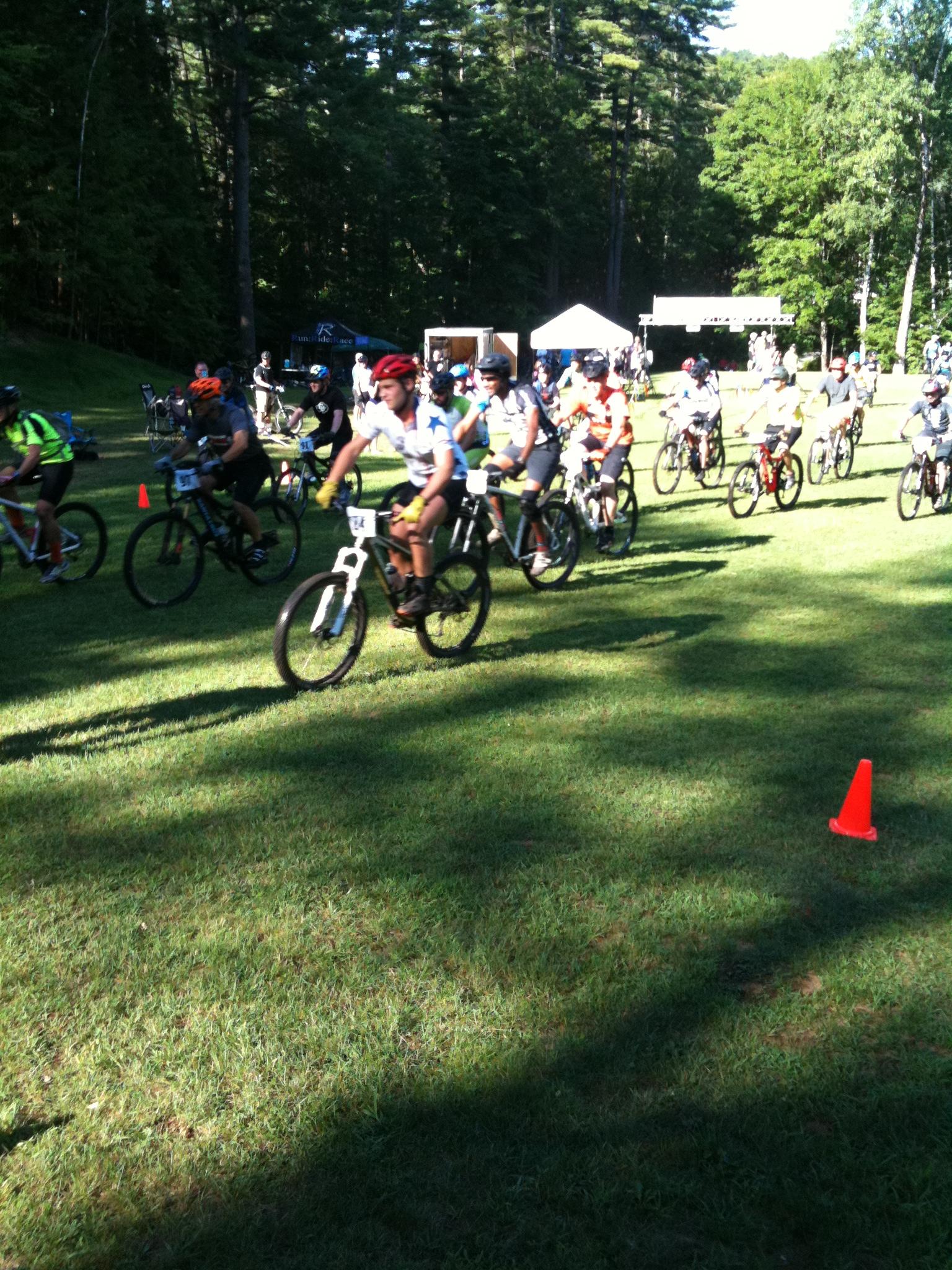 A group of mountain bikers, wearing helmets and colorful jerseys, are racing on a grassy field in a wooded area. Some participants are riding past orange traffic cones, while a few tents can be seen in the background. The scene captures the excitement of a cycling event in a sunny, natural setting. Gurney Lane Mountain Bike Trails mountain bike trail.