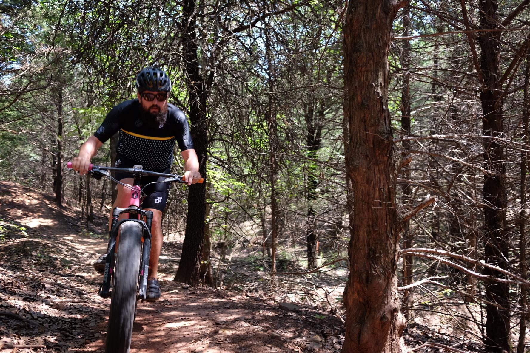 A mountain biker wearing a black and yellow cycling kit rides along a dirt trail in a wooded area, surrounded by trees and foliage. Sunlight filters through the leaves, creating a dappled light effect on the path. McMurtry Trail mountain bike trail.