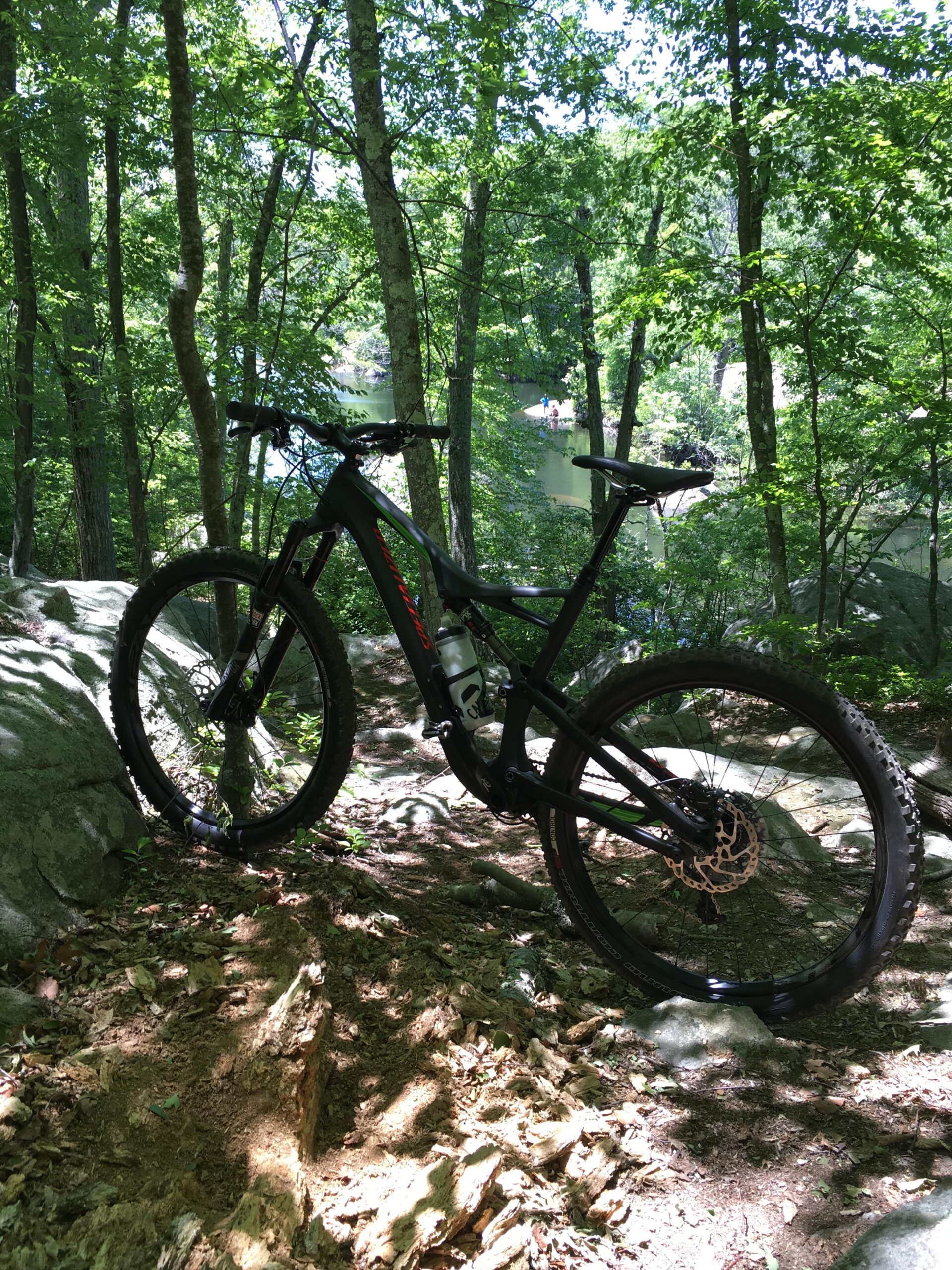 Specialized Camber 650B: A mountain bike resting on a rocky trail surrounded by lush green trees and foliage, with a glimpse of a river visible in the background. The bike features a black frame and is positioned on uneven terrain, showcasing its suitability for off-road rides.