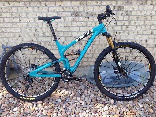 Yeti SB95: A blue mountain bike standing on a bed of gravel, with a brick wall in the background. The bike features thick tires, disc brakes, and a suspension fork.
