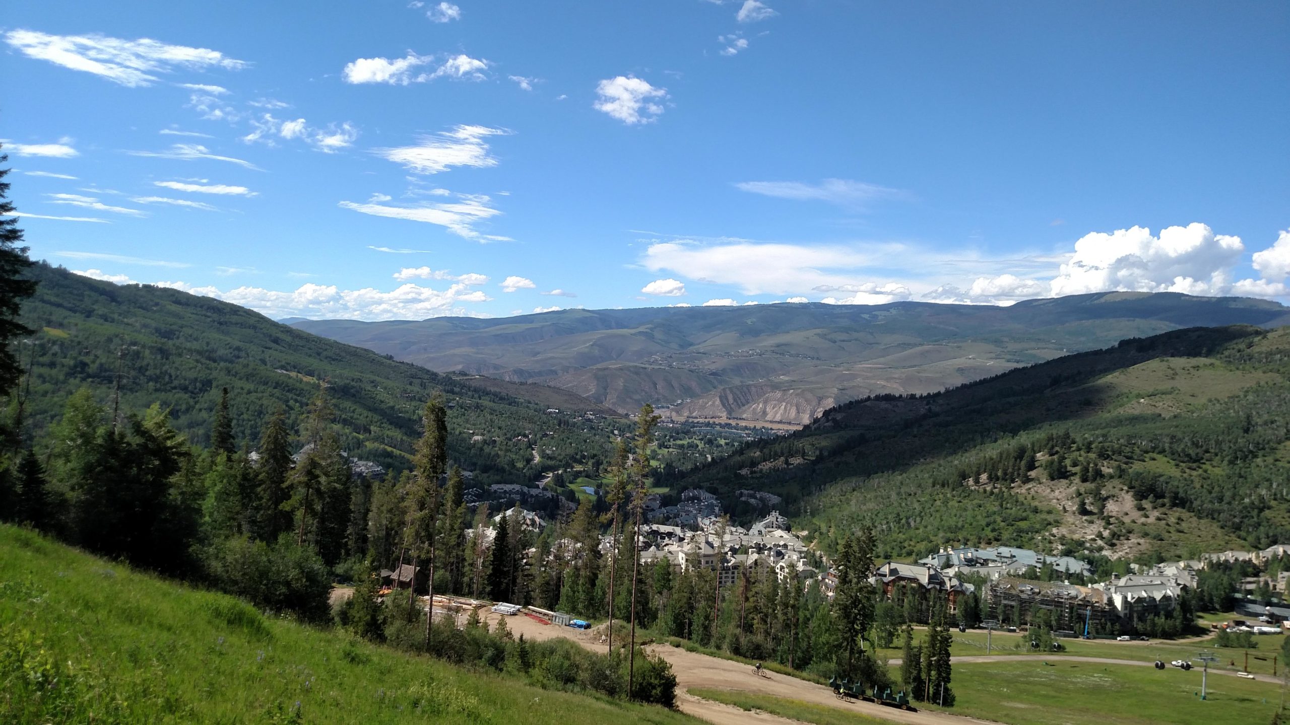 A scenic view of mountainous terrain with lush greenery and a small town nestled in the valley, under a bright blue sky with scattered clouds. The foreground features a mix of trees and grassy areas, while the background showcases rolling hills and distant peaks. Vail Mountain Bike Park mountain bike trail.
