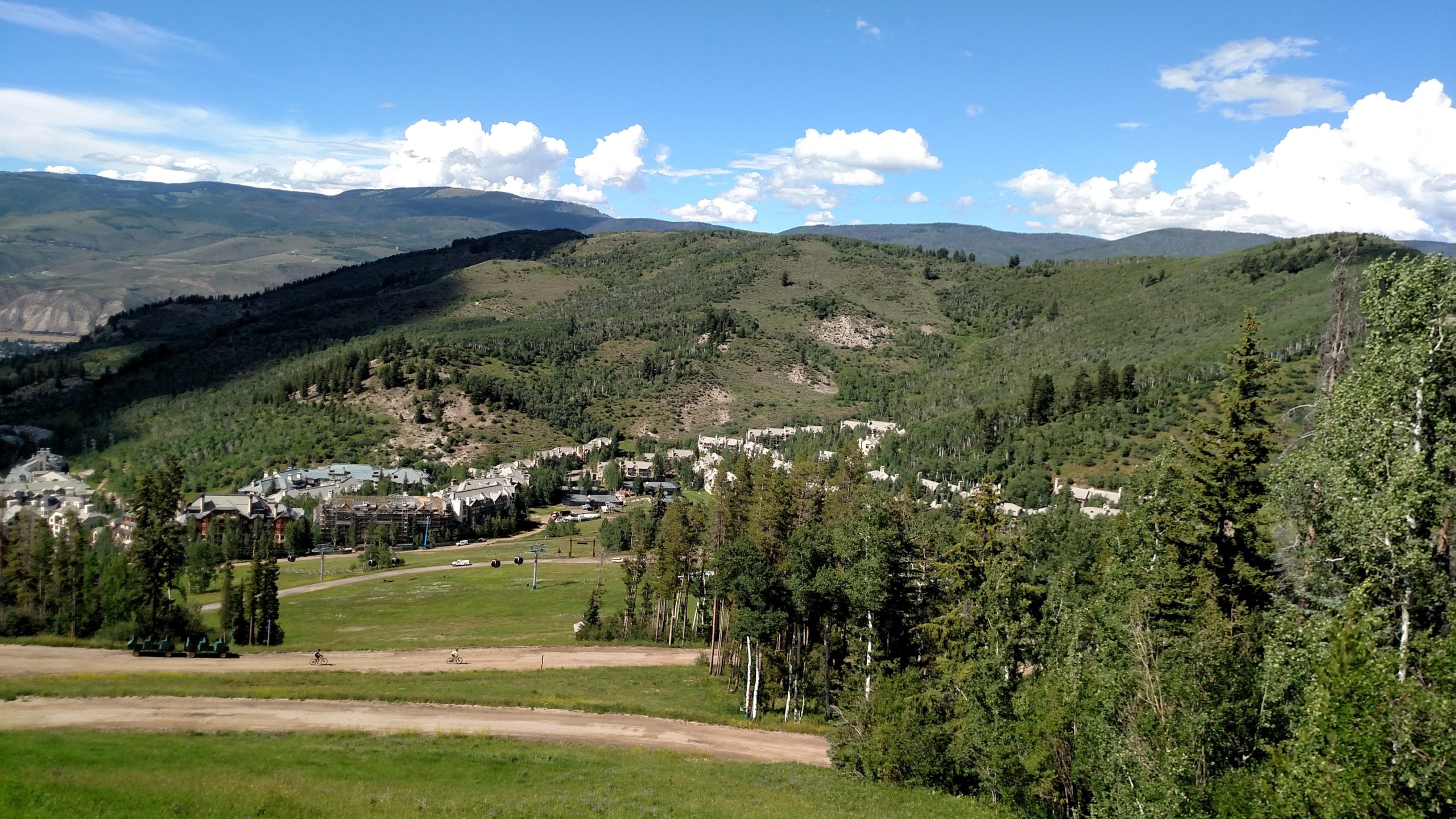 A scenic view of green mountains and valleys under a blue sky with fluffy white clouds. In the foreground, a grassy area with a few trees and a dirt path, while in the midground, a small town or resort area is nestled among the trees. The landscape showcases various shades of green and hints of development, with a few visible buildings and structures. Vail Mountain Bike Park mountain bike trail.