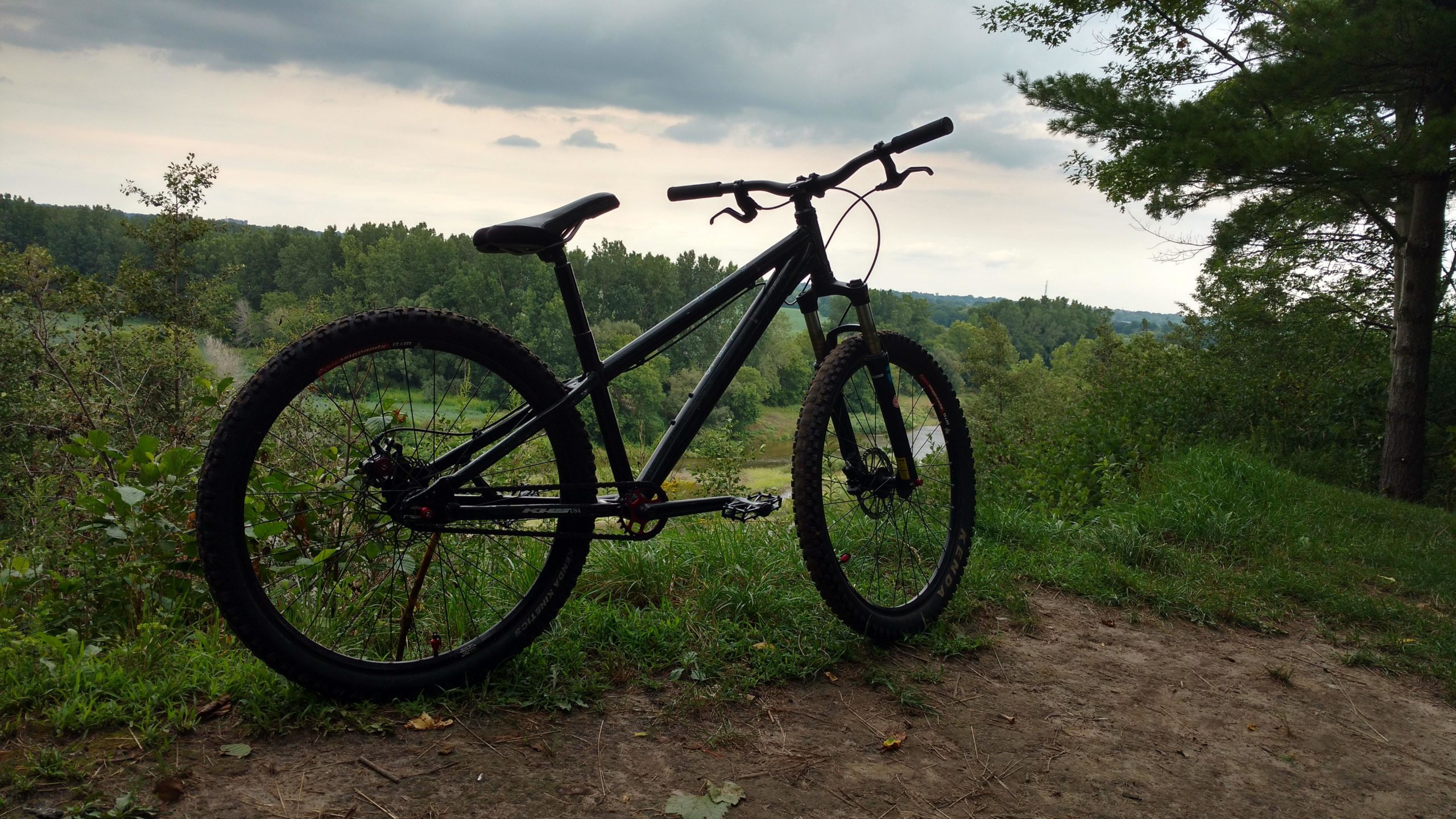 A black mountain bike is positioned on a dirt path overlooking a lush green landscape with trees and rolling hills. The sky is overcast with a few clouds. The bike