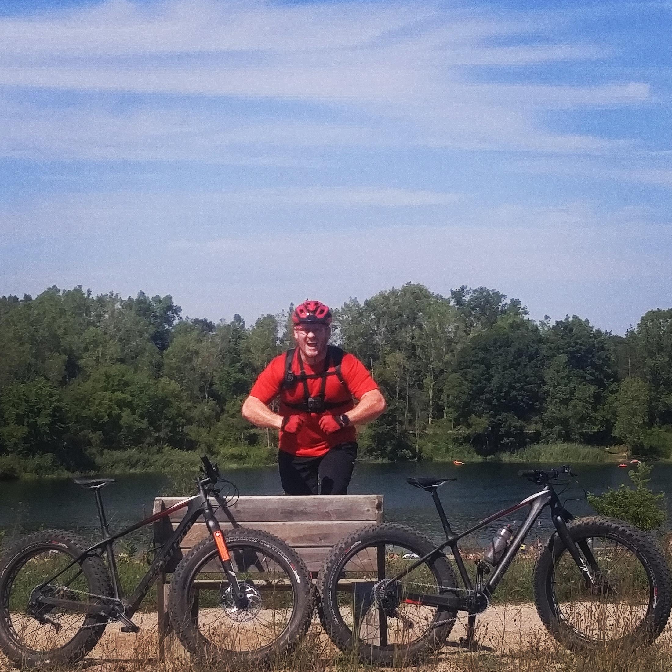 A person wearing a red helmet and shirt stands next to two mountain bikes parked on a wooden bench near a scenic lake. The background features lush greenery and a clear blue sky. Island Lake mountain bike trail.