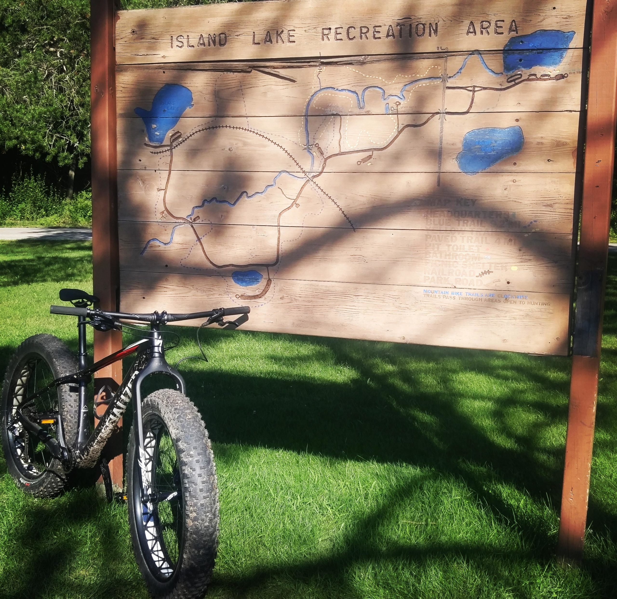 A fat tire bike parked next to a wooden sign displaying a map of the Island Lake Recreation Area, with blue markings indicating trails and bodies of water. The surrounding area features green grass and trees, suggesting a natural outdoor setting. Island Lake mountain bike trail.