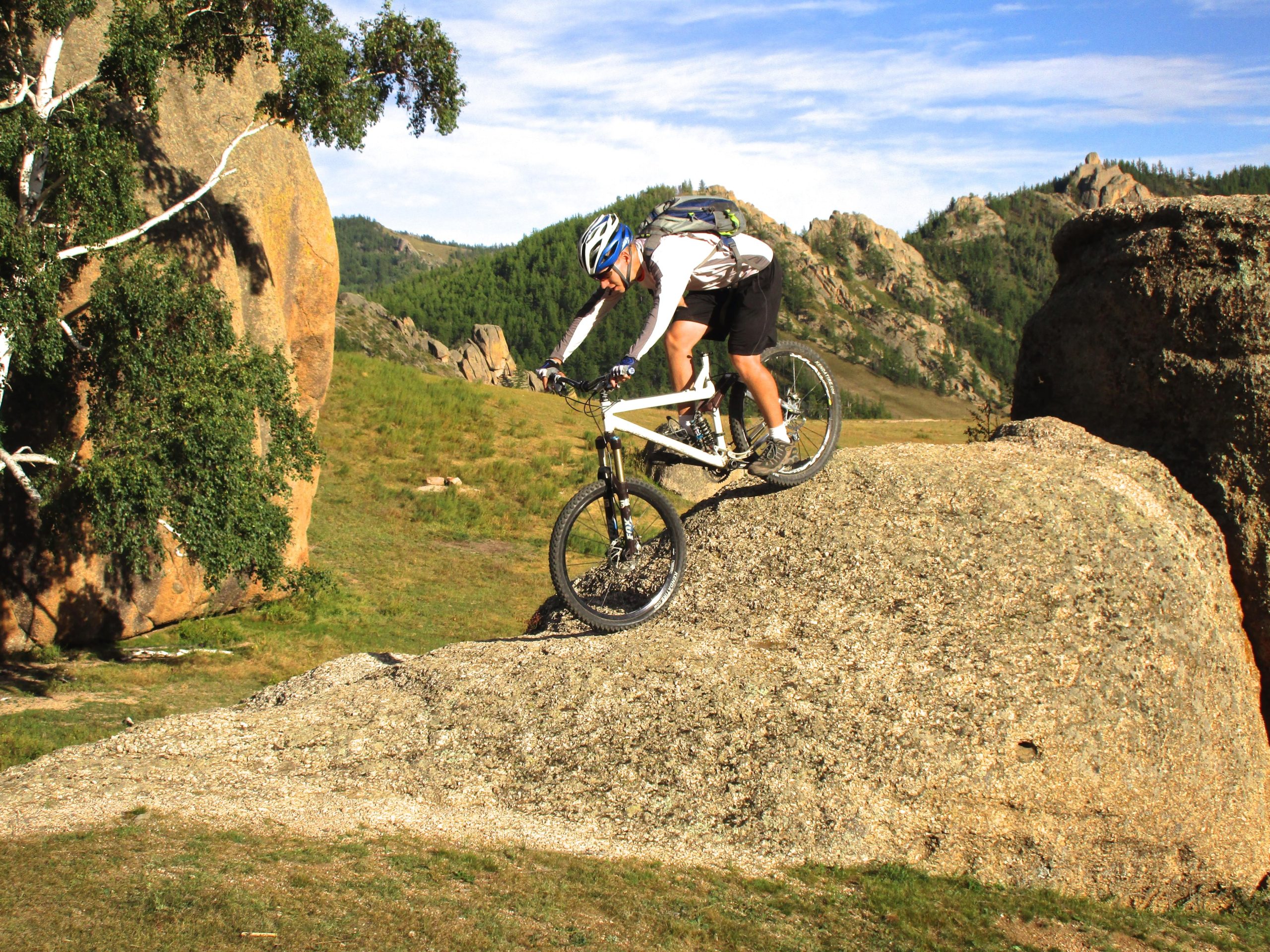 A mountain biker skillfully descends a large rock in a scenic outdoor landscape, surrounded by greenery and rugged terrain. The rider is wearing a helmet and cycling gear, focusing on maintaining balance while navigating the rocky surface. Camel Run mountain bike trail.