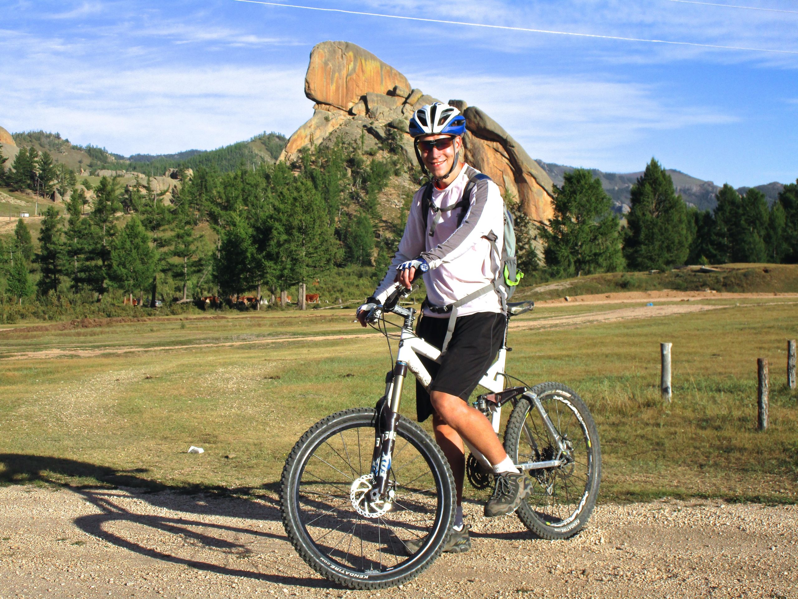 A smiling cyclist wearing a helmet and sunglasses is posed on a mountain bike, set against a backdrop of rocky hills and green trees. The scene captures a sunny day with clear skies, showcasing a sense of adventure and the beauty of the outdoors. Camel Run mountain bike trail.