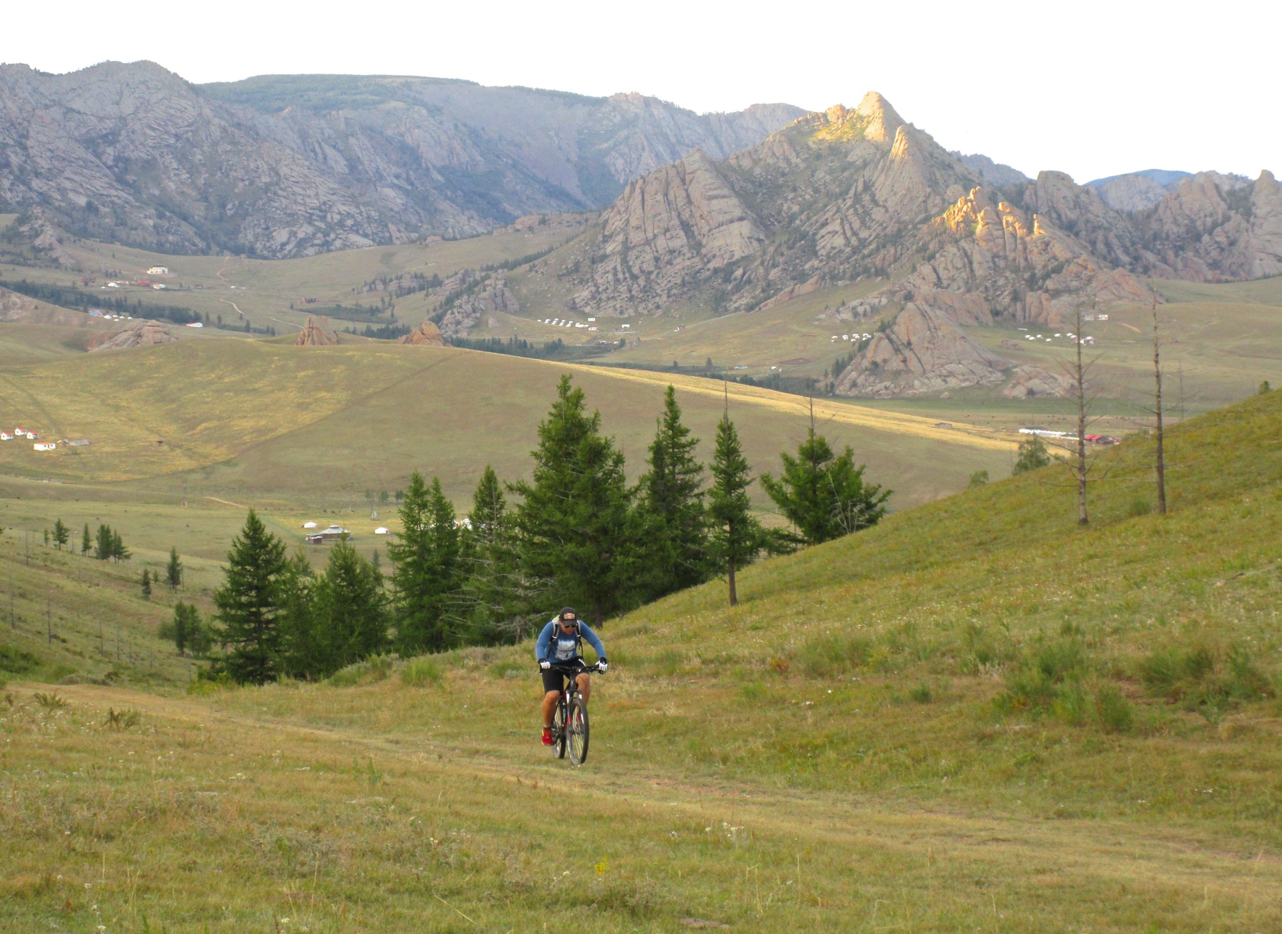 A mountain biker riding uphill on a grassy slope, surrounded by trees and mountains in the background. The landscape features rolling hills and rocky formations with distant buildings visible. The scene is illuminated by soft sunlight, creating a serene outdoor atmosphere. Camel Run mountain bike trail.