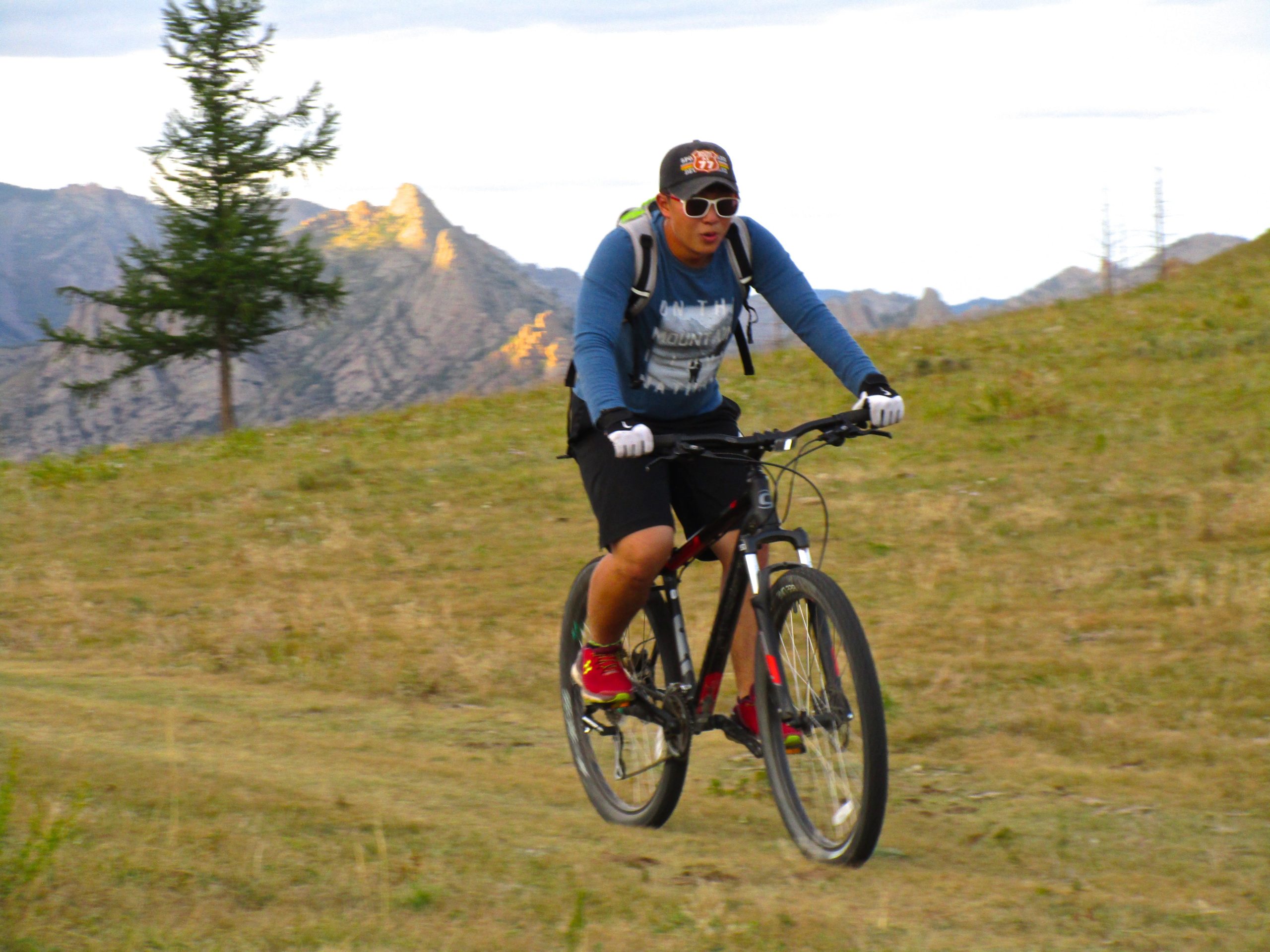 A person riding a mountain bike on a grassy trail, surrounded by mountainous terrain in the background. The rider is wearing a blue long-sleeve shirt, black shorts, and sunglasses, with a backpack and gloves. Trees and rocky peaks are visible in the landscape under a partly cloudy sky. Camel Run mountain bike trail.