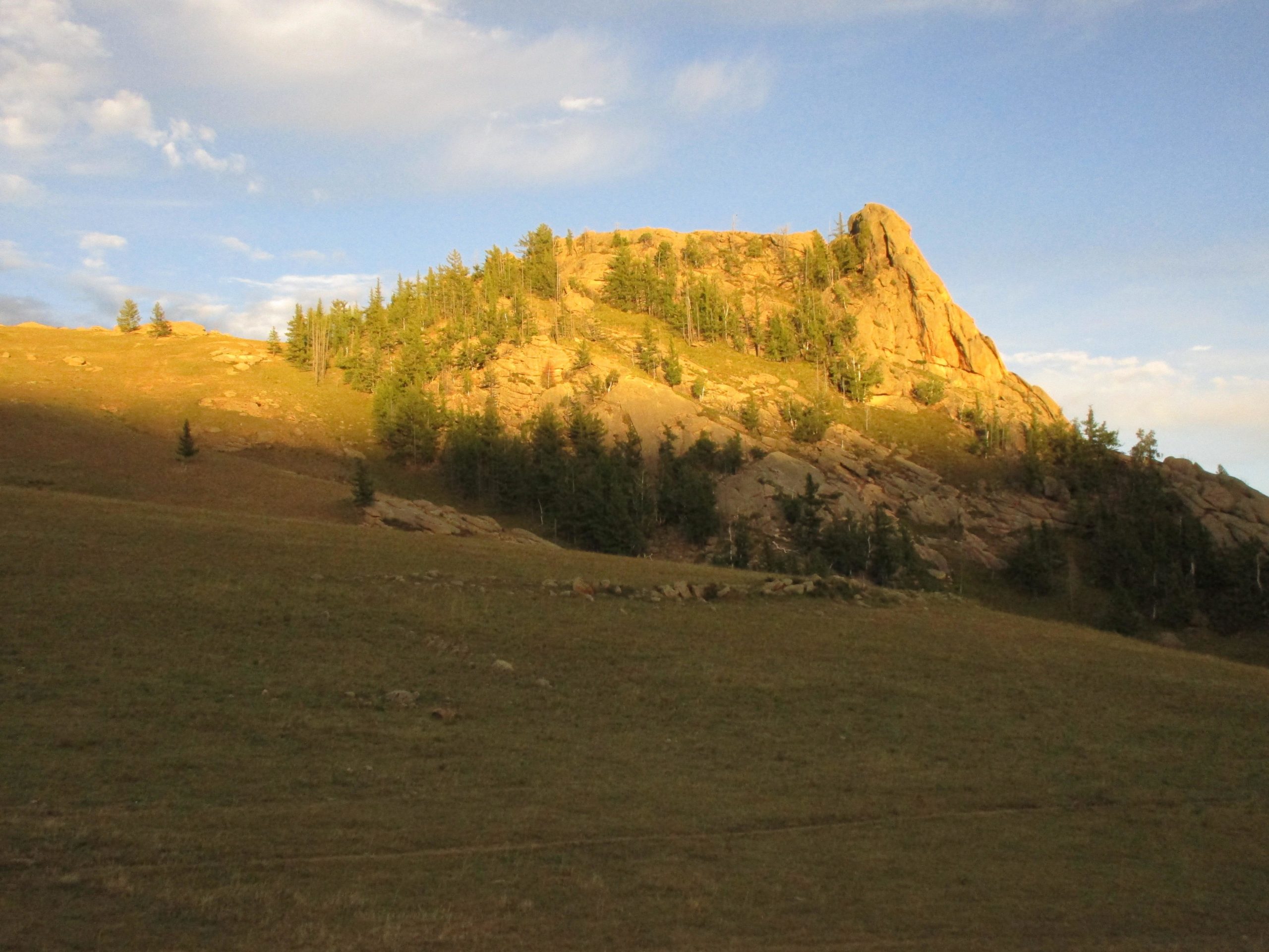 A rugged rock formation illuminated by sunlight, set against a blue sky with scattered clouds. Lush green trees grow on the slope, surrounded by a grassy landscape. The scene conveys a serene natural environment. Camel Run mountain bike trail.
