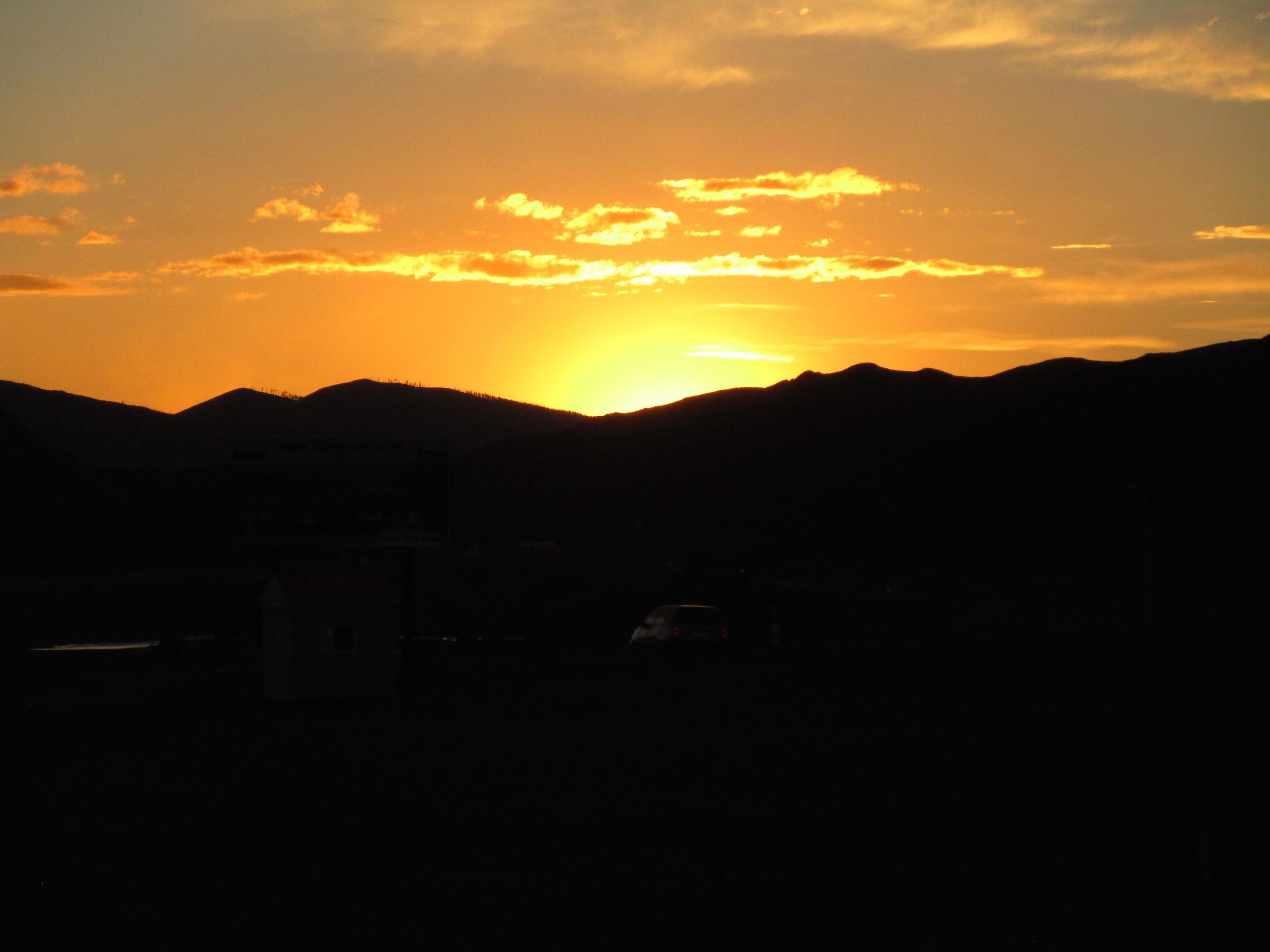 Sunset over silhouetted mountains, with vibrant orange and yellow hues illuminating the sky and casting a warm glow. A small building and vehicles are visible in the foreground, creating a serene landscape scene. Camel Run mountain bike trail.