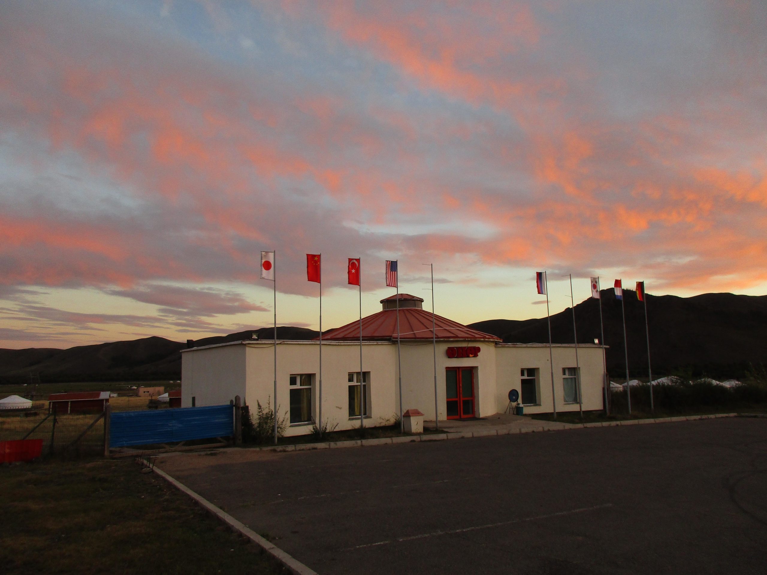 A white building with a red roof, prominently displaying several flags from different countries, under a colorful sky at sunset. The surrounding landscape includes rolling hills and sparse vegetation. Camel Run mountain bike trail.