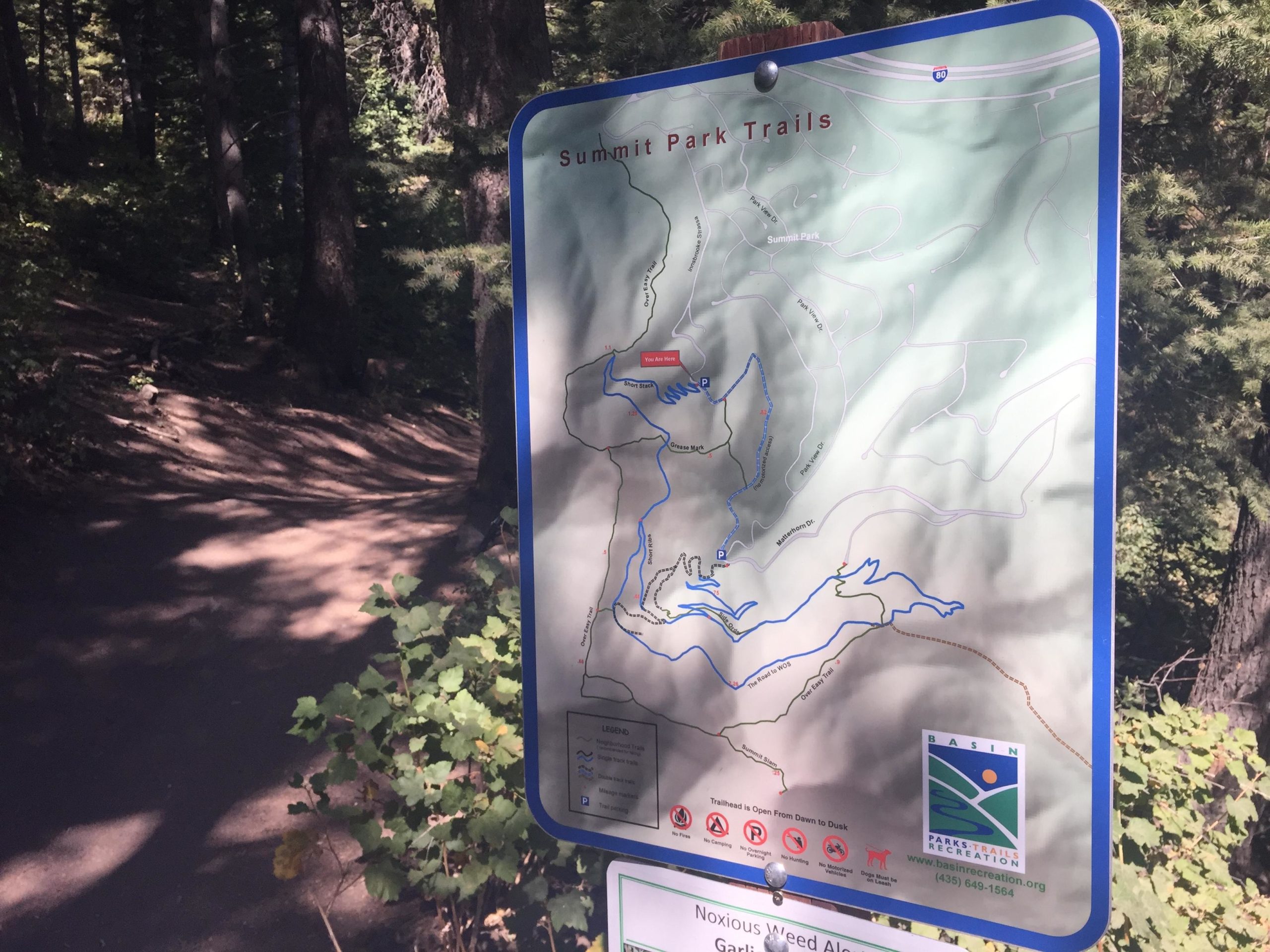 Map of Summit Park Trails indicating various hiking routes, with details on trailheads and terrain. The sign is positioned near a dirt path surrounded by trees. The Road to WOS mountain bike trail.