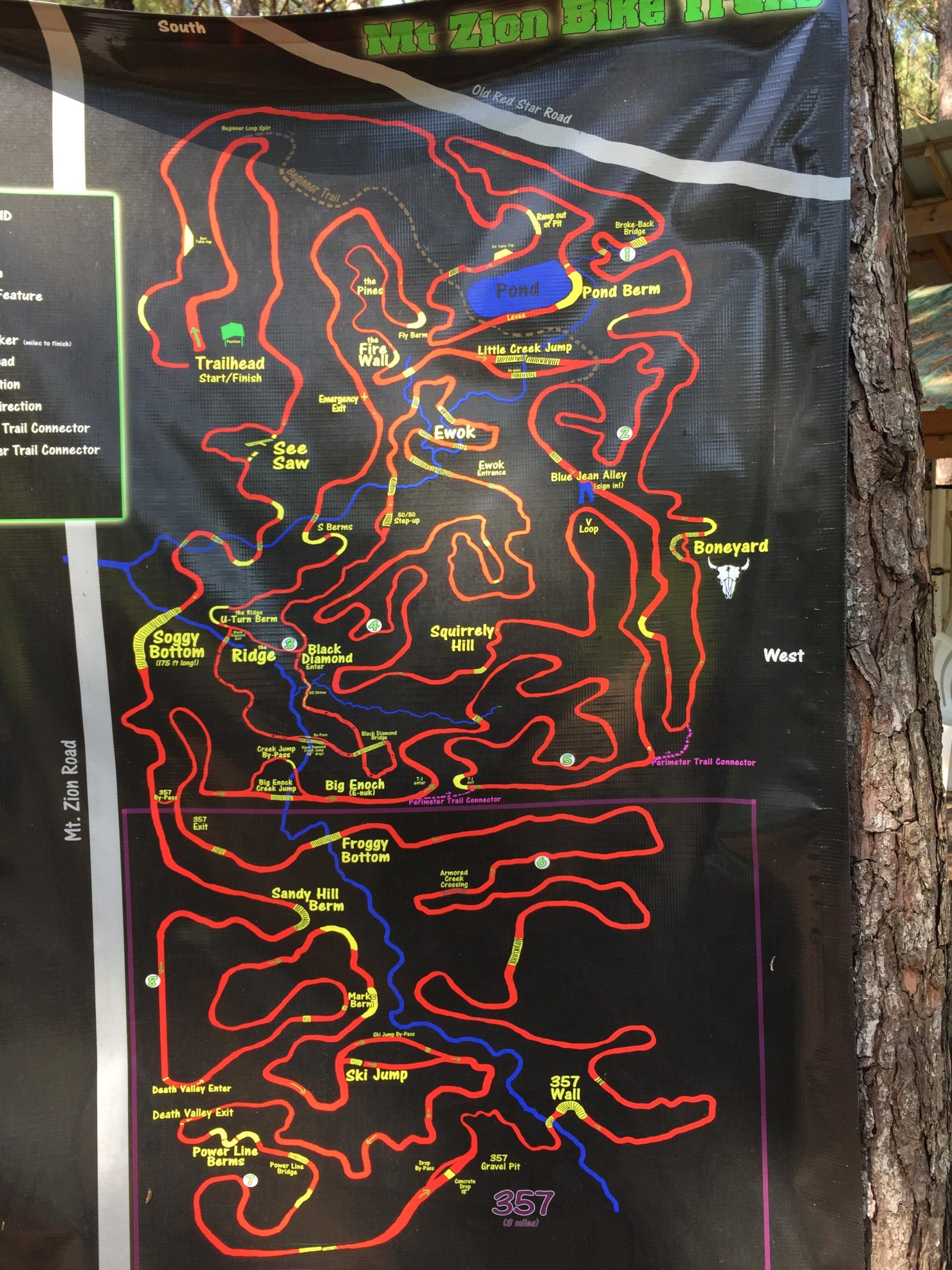 A detailed map of the Mt. Zion Bike Trails, featuring various marked routes in red and blue, along with key locations such as the trailhead, pond, and different trail features. The map indicates multiple trail names, including "Ewok," "Sandy Hill Berm," and "357 Wall," and highlights directional paths and important connectors. Mt. Zion Bike Trails mountain bike trail.