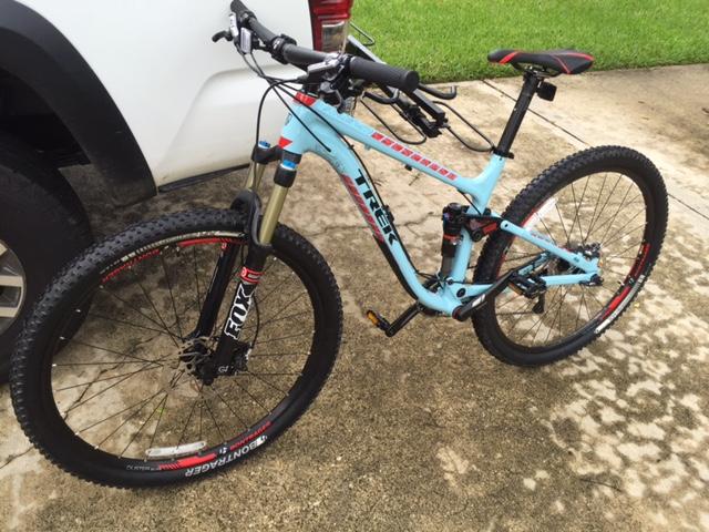 Trek Fuel EX 8 29: A light blue Trek mountain bike parked next to a vehicle, featuring a front suspension fork, thick tires, and red-accented rims. The bike is positioned on a concrete surface with grass in the background.